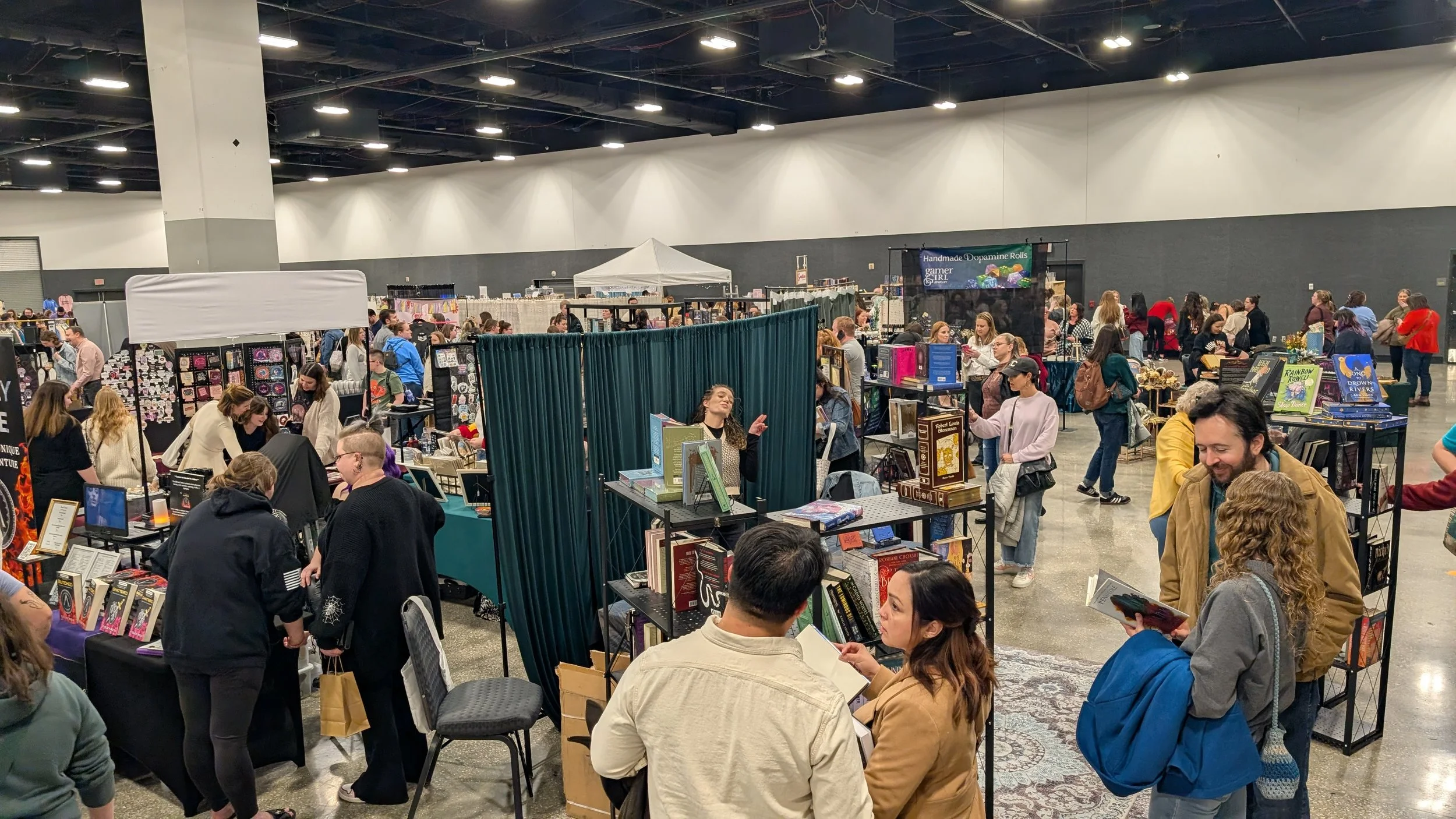 Crowd of people browsing vendors at a convention or trade show, with tables displaying books and merchandise.