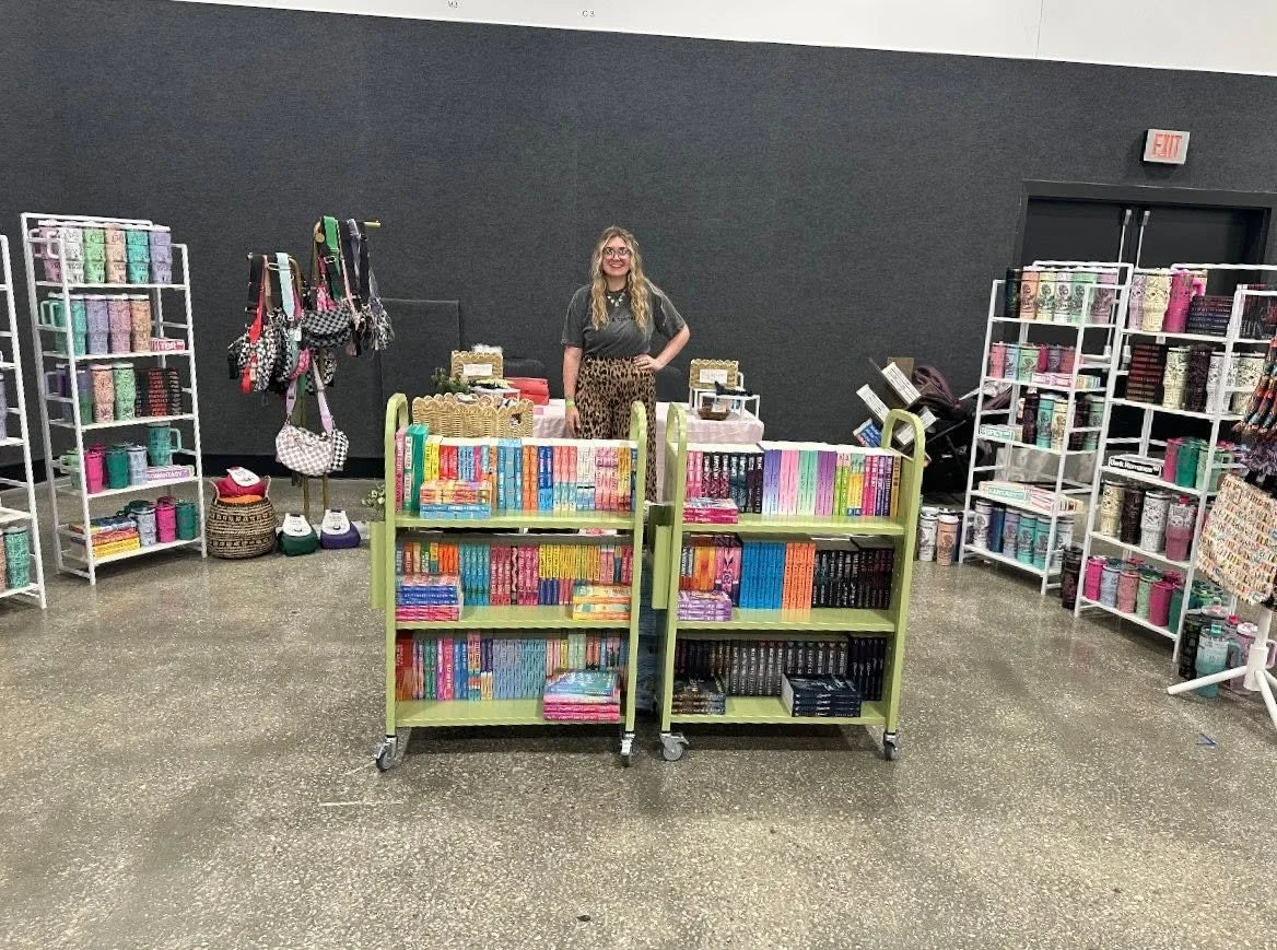 A woman standing behind a table at a bookstore or craft fair, surrounded by shelves of colorful books and patterned bags.