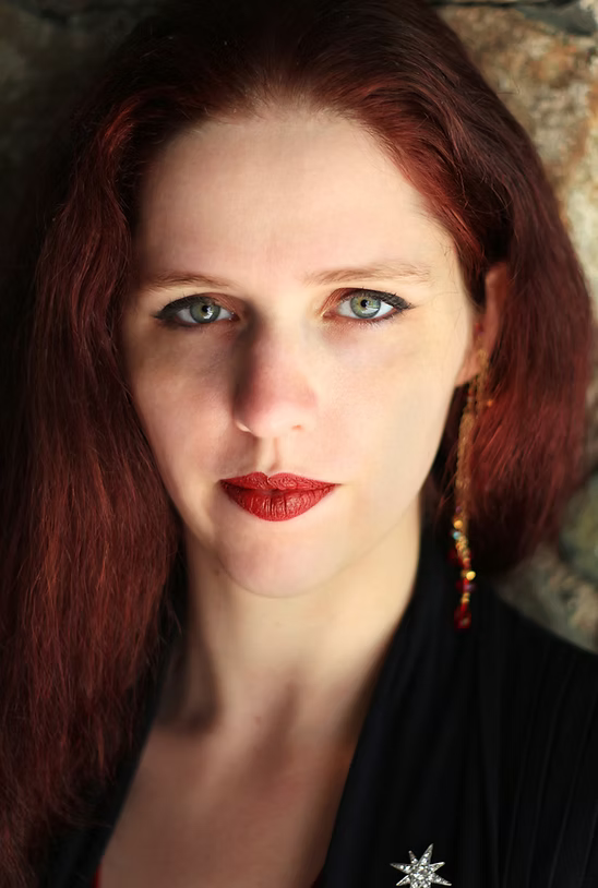 A woman with long red hair, blue eyes, and red lipstick, wearing a black top and a colorful earring, standing outdoors near a stone wall.