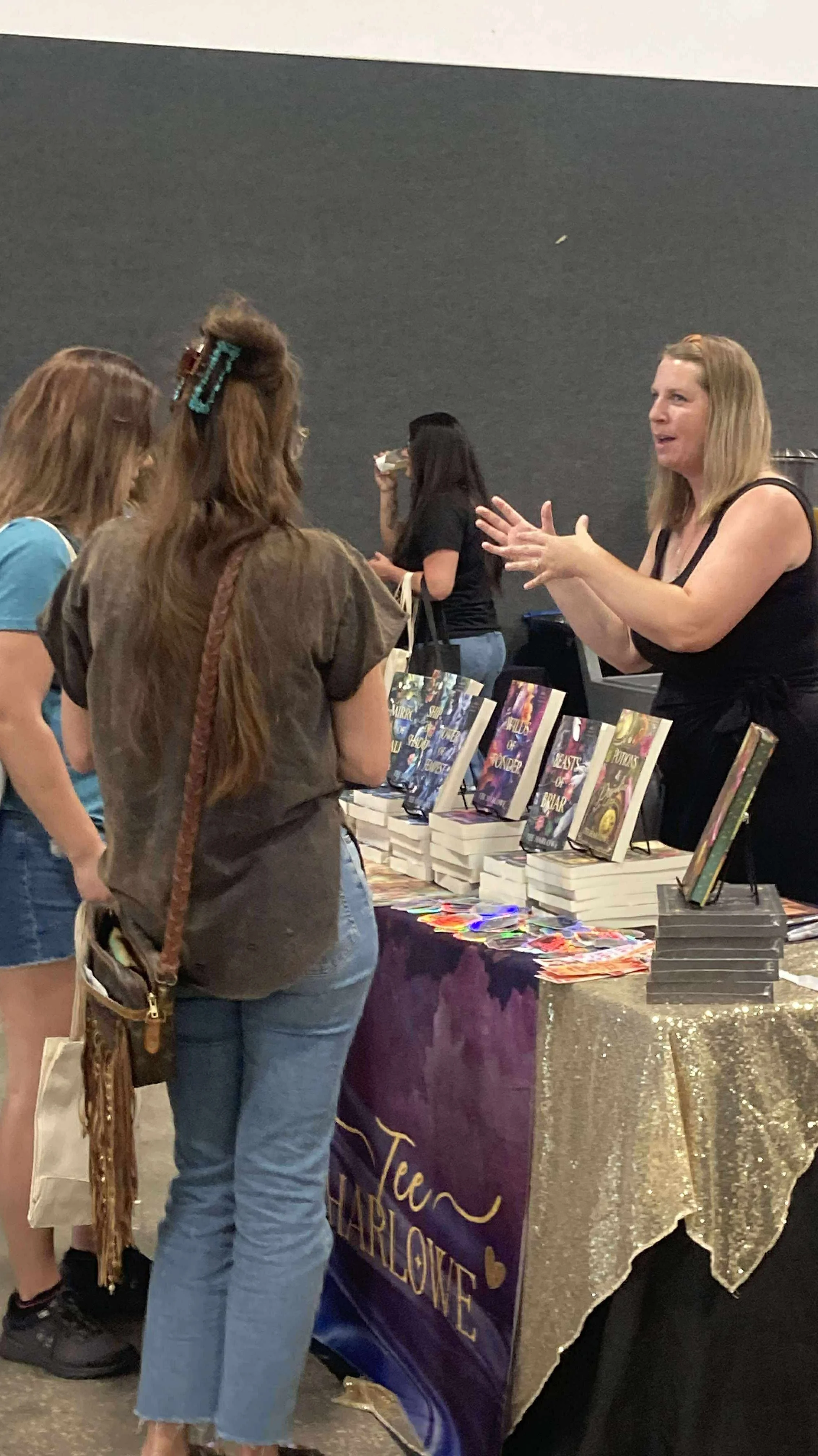 Women browsing books at a vendor table with a purple and gold decorated tablecloth, books on display, and a woman behind the table talking to a customer, at an indoor event.