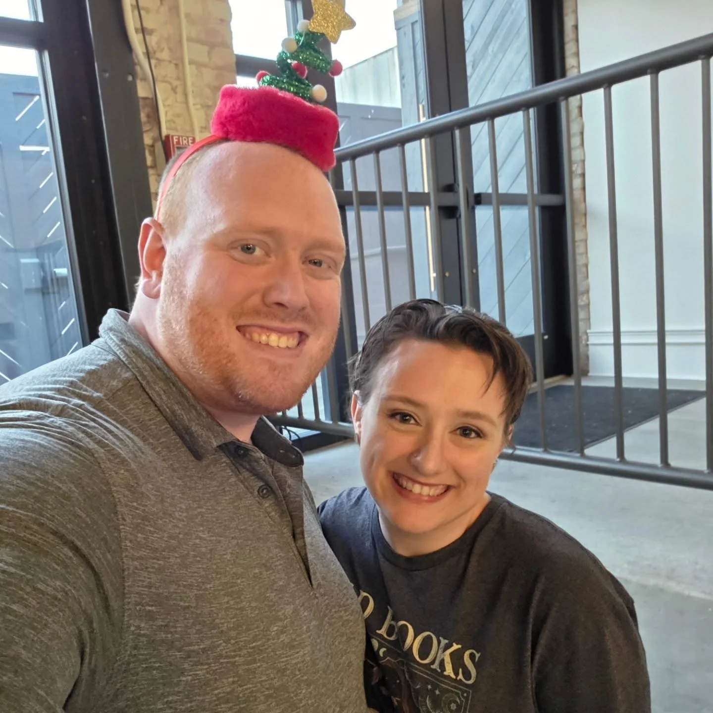 Two smiling people taking a selfie indoors, with a small Christmas tree headband worn by the person on the left, and a festive black t-shirt worn by the person on the right.