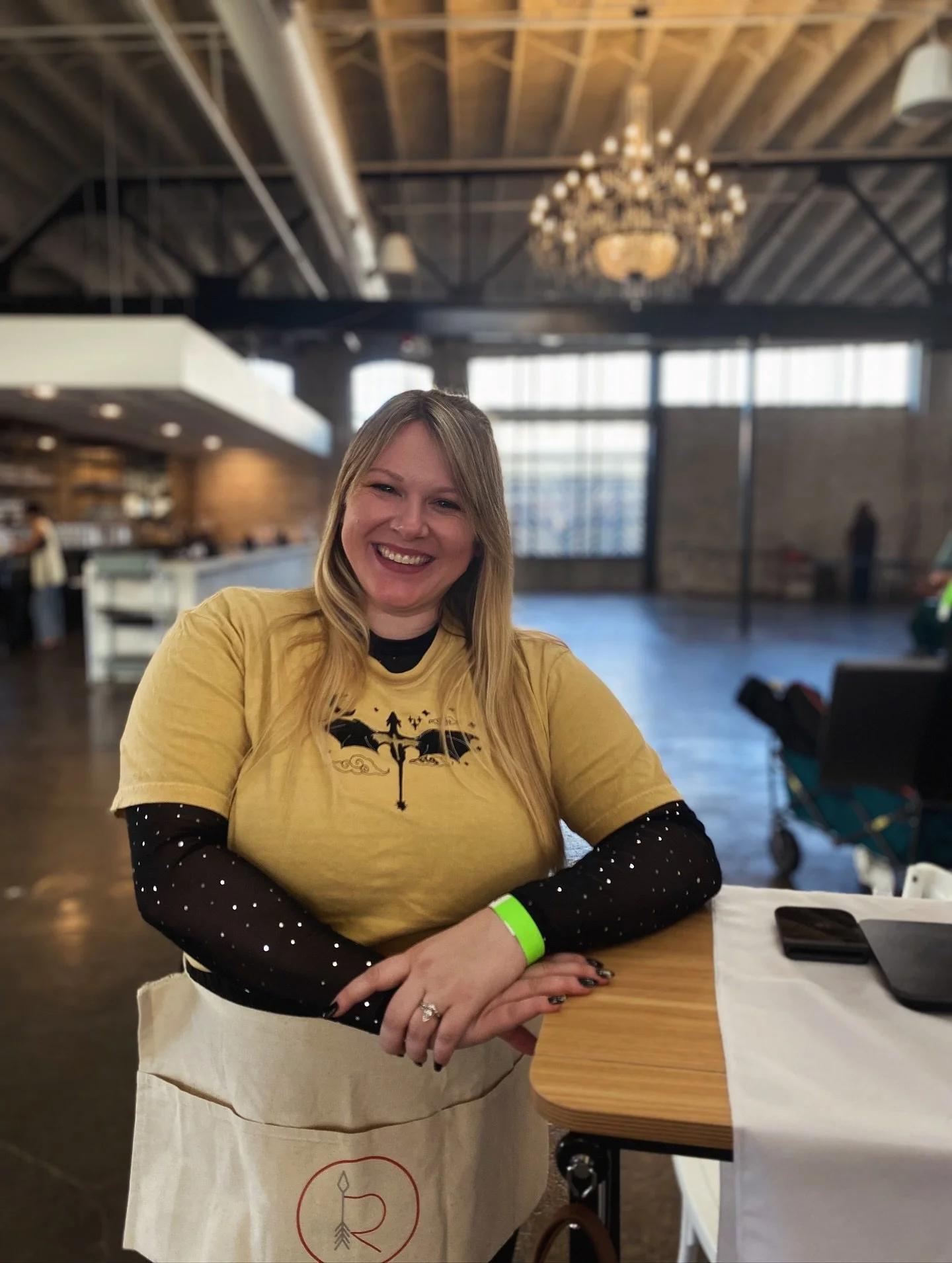 A woman with long blonde hair smiling, sitting at a table in a modern, industrial-style restaurant with a high ceiling, exposed ductwork, and a large chandelier. She is wearing a yellow T-shirt with a black graphic and a black, polka-dotted long-slee