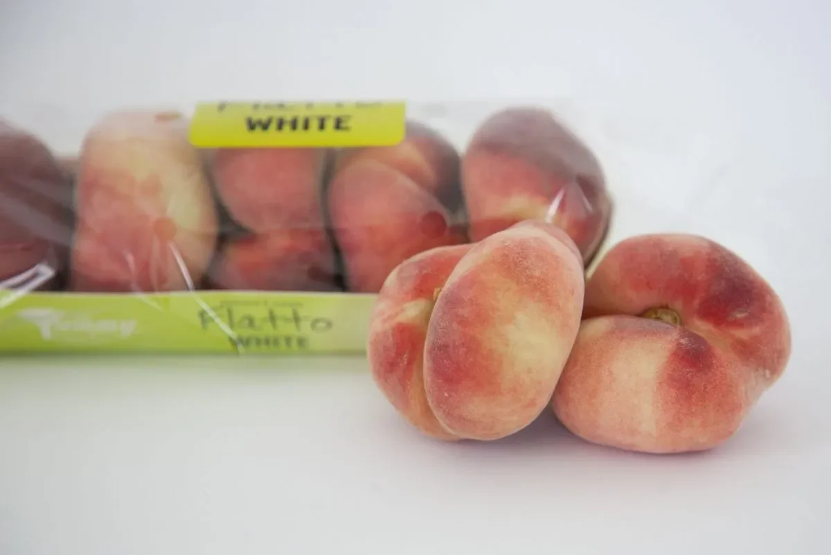 A close-up of fresh peaches with fuzzy skin beside a plastic container of peaches labeled 'NATTO WHITE' on a white background.