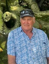 A man in a blue patterned shirt and sunglasses standing outdoors near a fallen tree with a large panda resting on it.