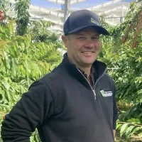 Smiling man wearing a black jacket and baseball cap standing in a greenhouse with green plants.