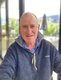 A smiling middle-aged man with short light hair, wearing a blue sweater and a checkered shirt, sitting indoors with a window view of trees and outdoor umbrellas behind him.