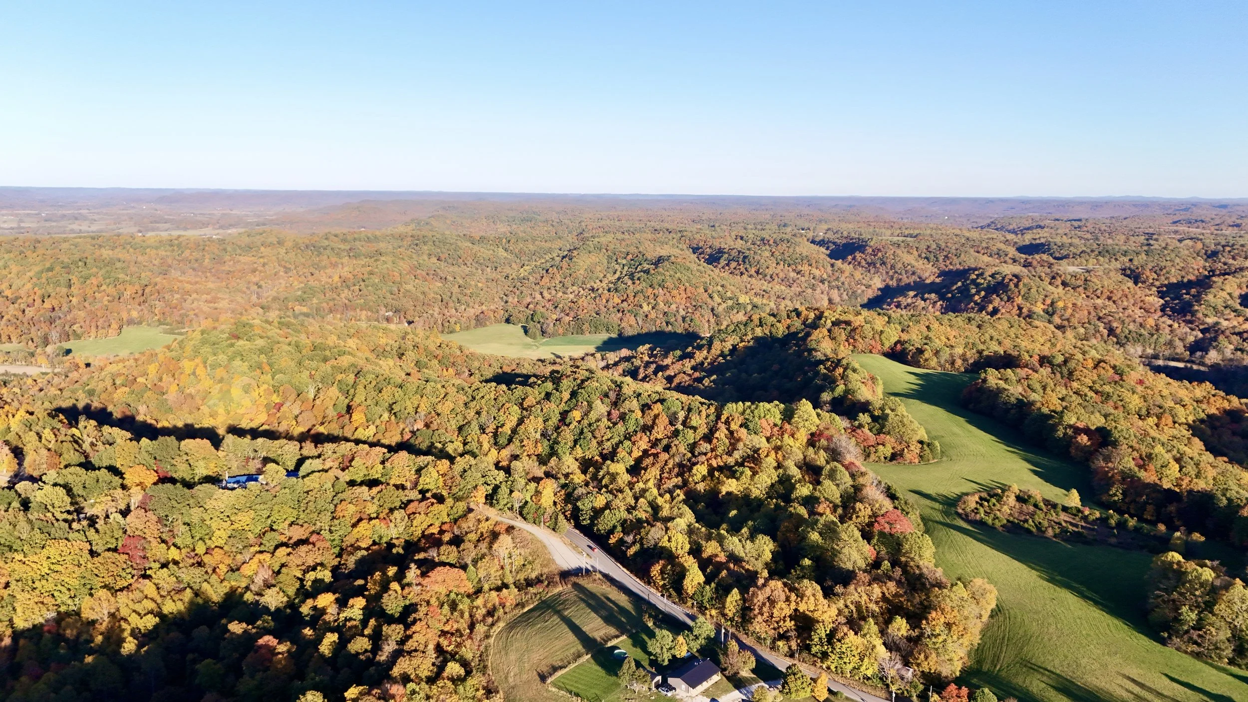 Aerial view of rolling hills covered in colorful autumn foliage, with green fields, a winding road, and a few buildings under a clear blue sky.