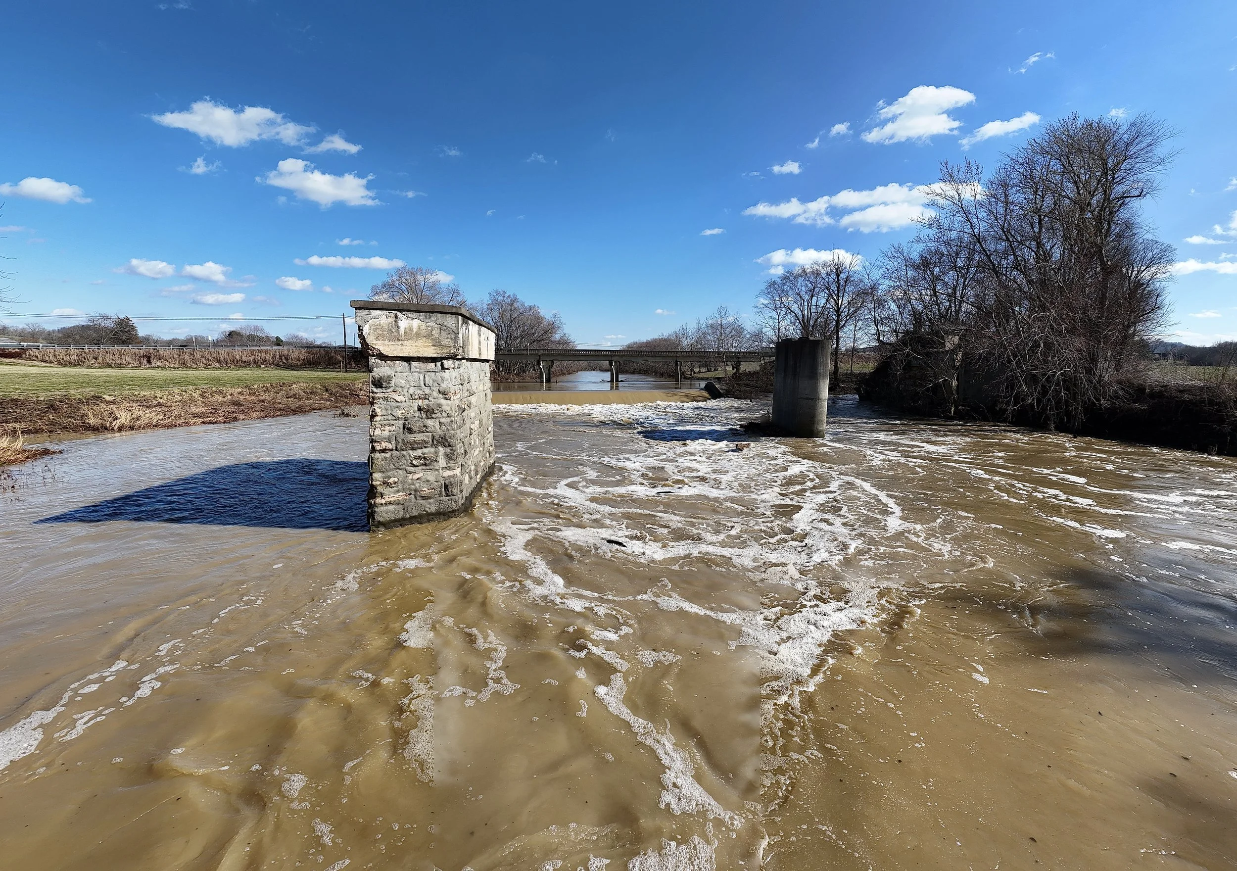 Flooded river with muddy water, stone pillars, and a bridge in the background under a bright blue sky with scattered clouds.