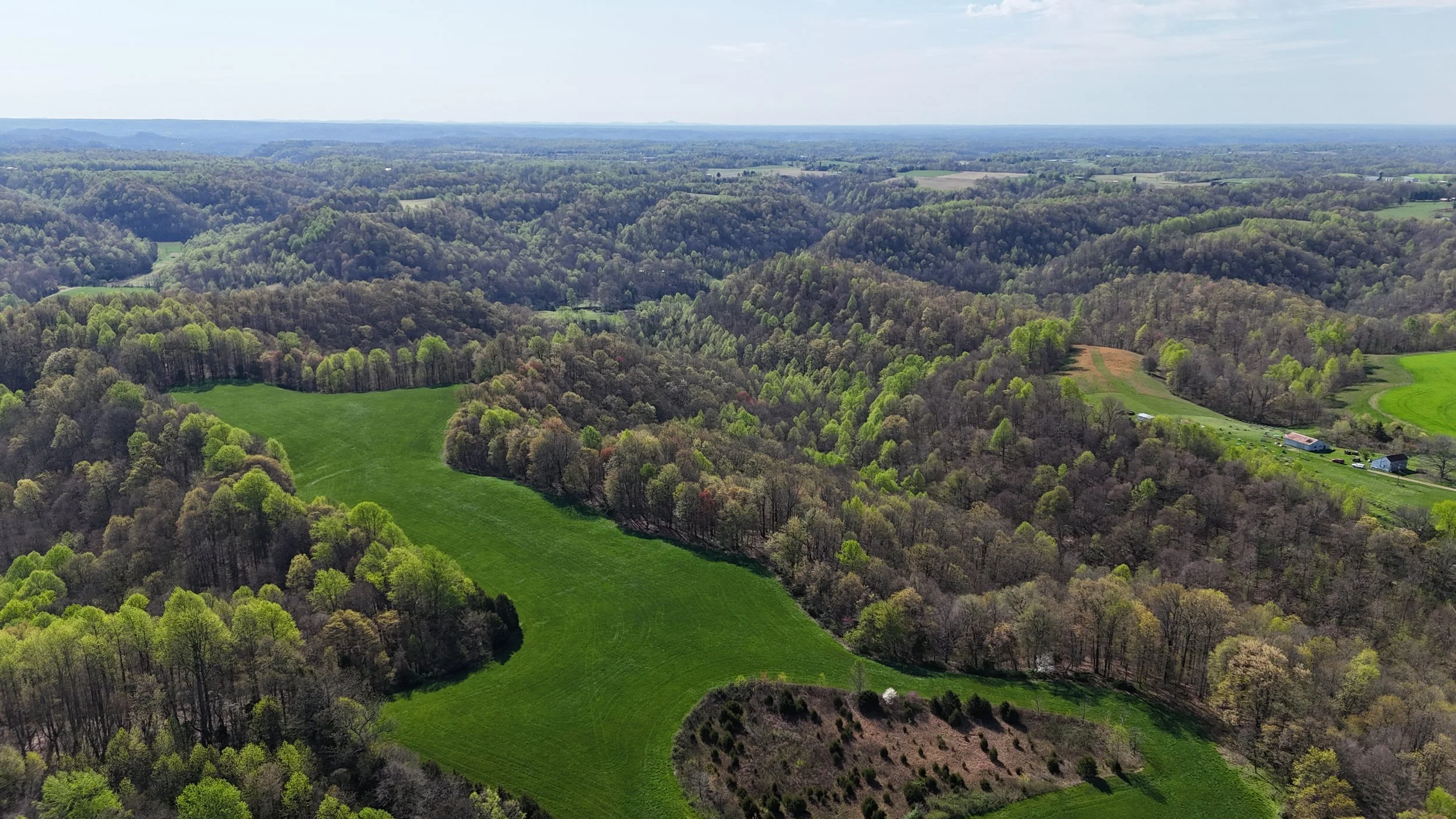 Aerial view of a lush green landscape with rolling hills, dense forests, and farm buildings in a rural area.