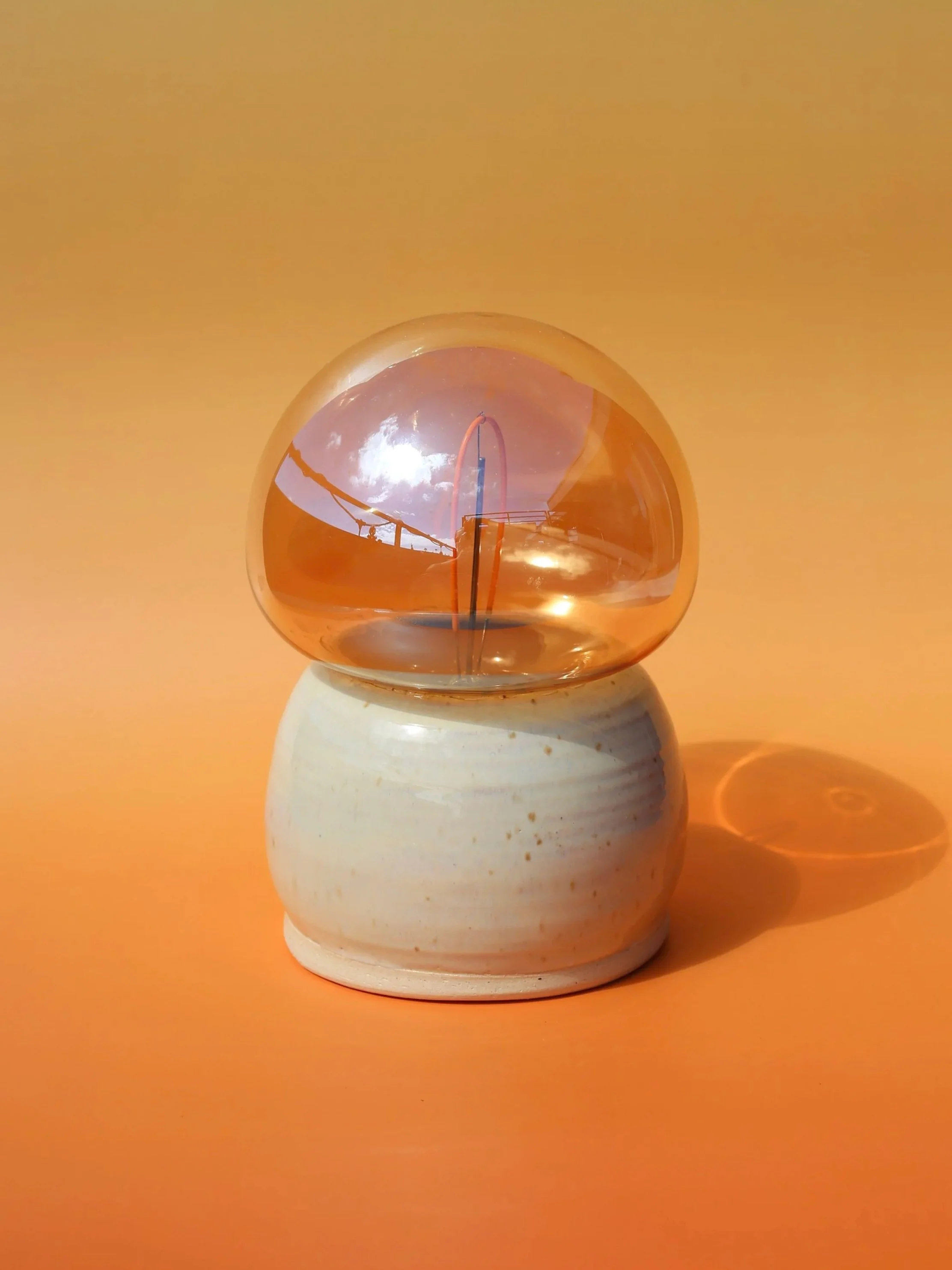 A small spherical orange lamp with a warm clear mushroom shaped lightbulb, placed on a handmade white sandy beige ceramic vase against an orange background.
