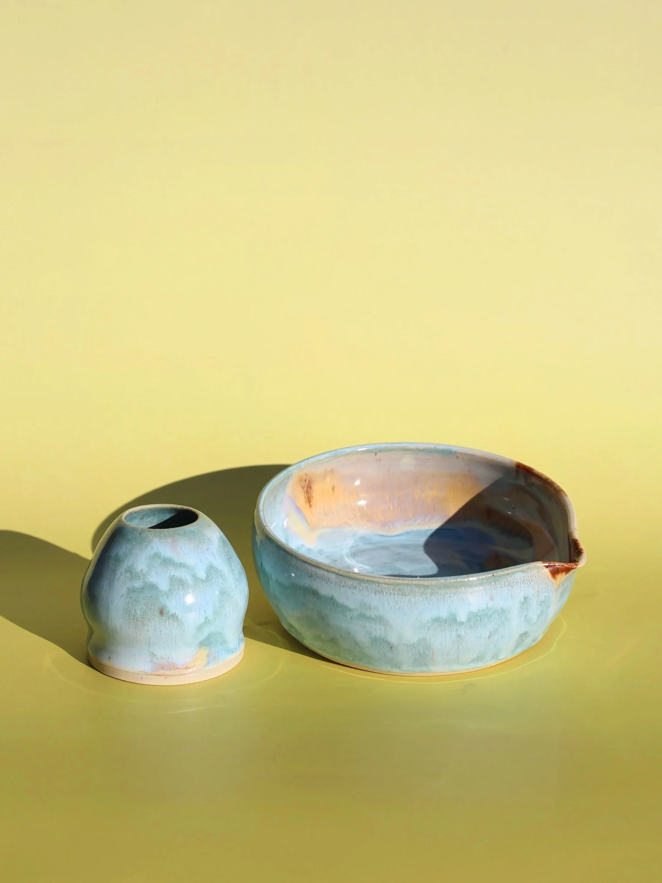 Handmade ceramic matcha bowl and matcha whisk chasen holder set with a glossy, pale blue glaze, set against a yellow background.