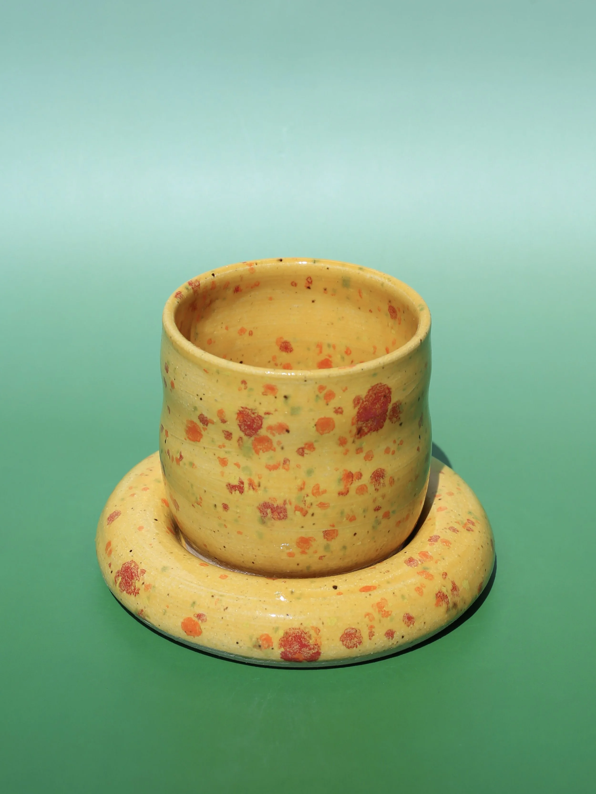 A yellow ceramic vase with orange, green, and black speckles, placed on a matching yellow ceramic plate against a light blue background.