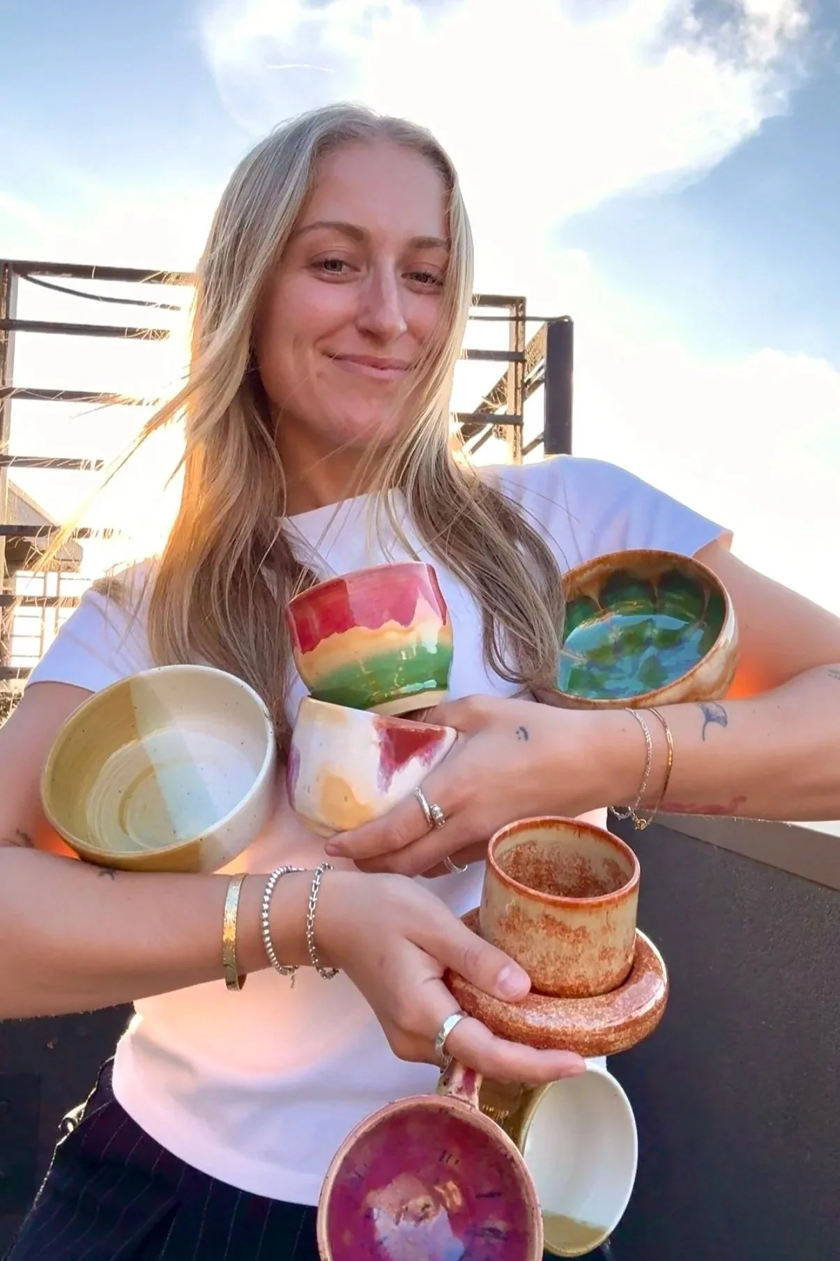 A woman with long blonde hair holding multiple colorful handmade ceramic bowls and cups, standing outdoors against a partly cloudy sky.