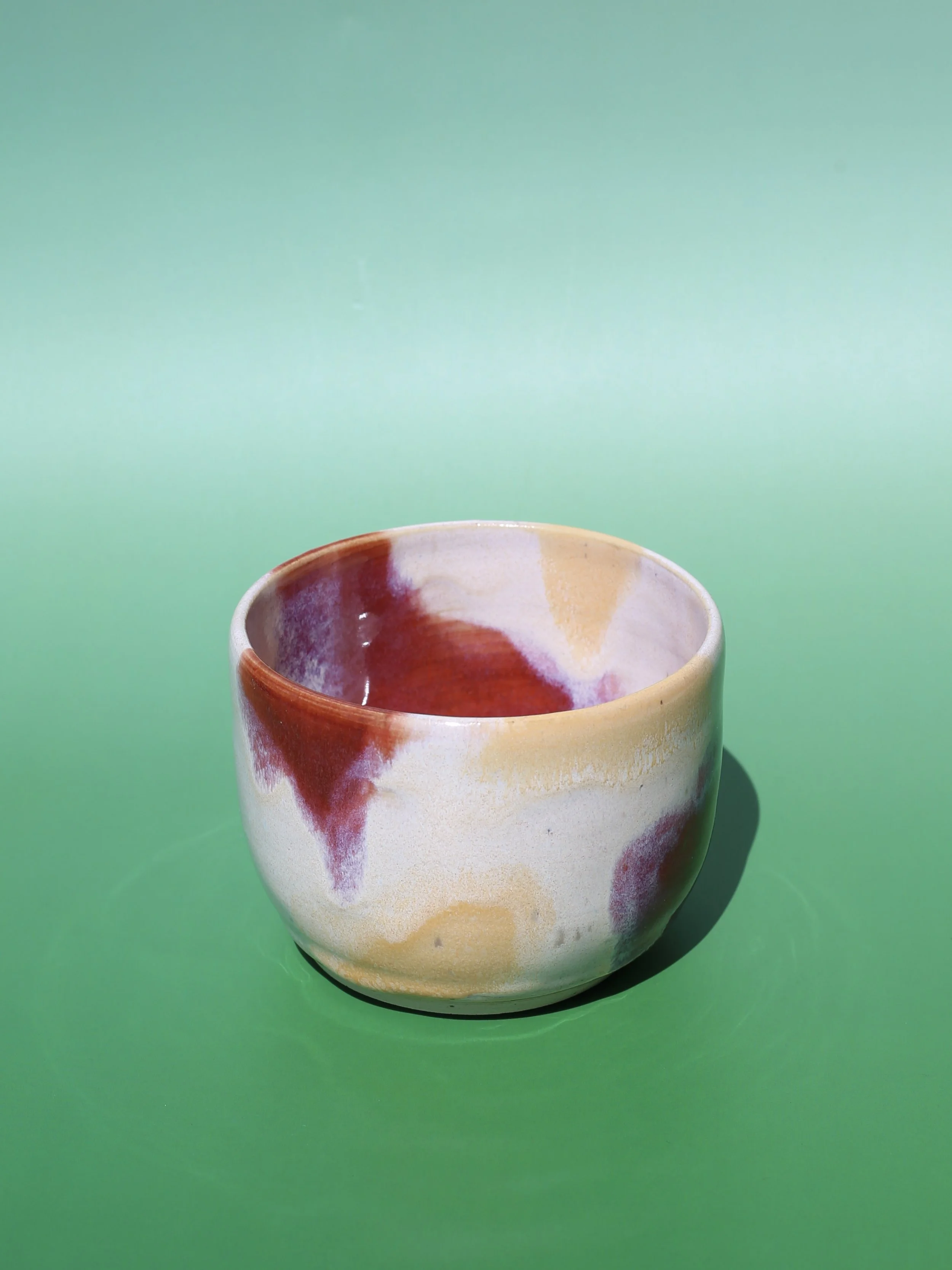 A ceramic cup with a colorful, abstract glaze design in shades of red, beige, and cream, sitting on a green surface against a plain green background.