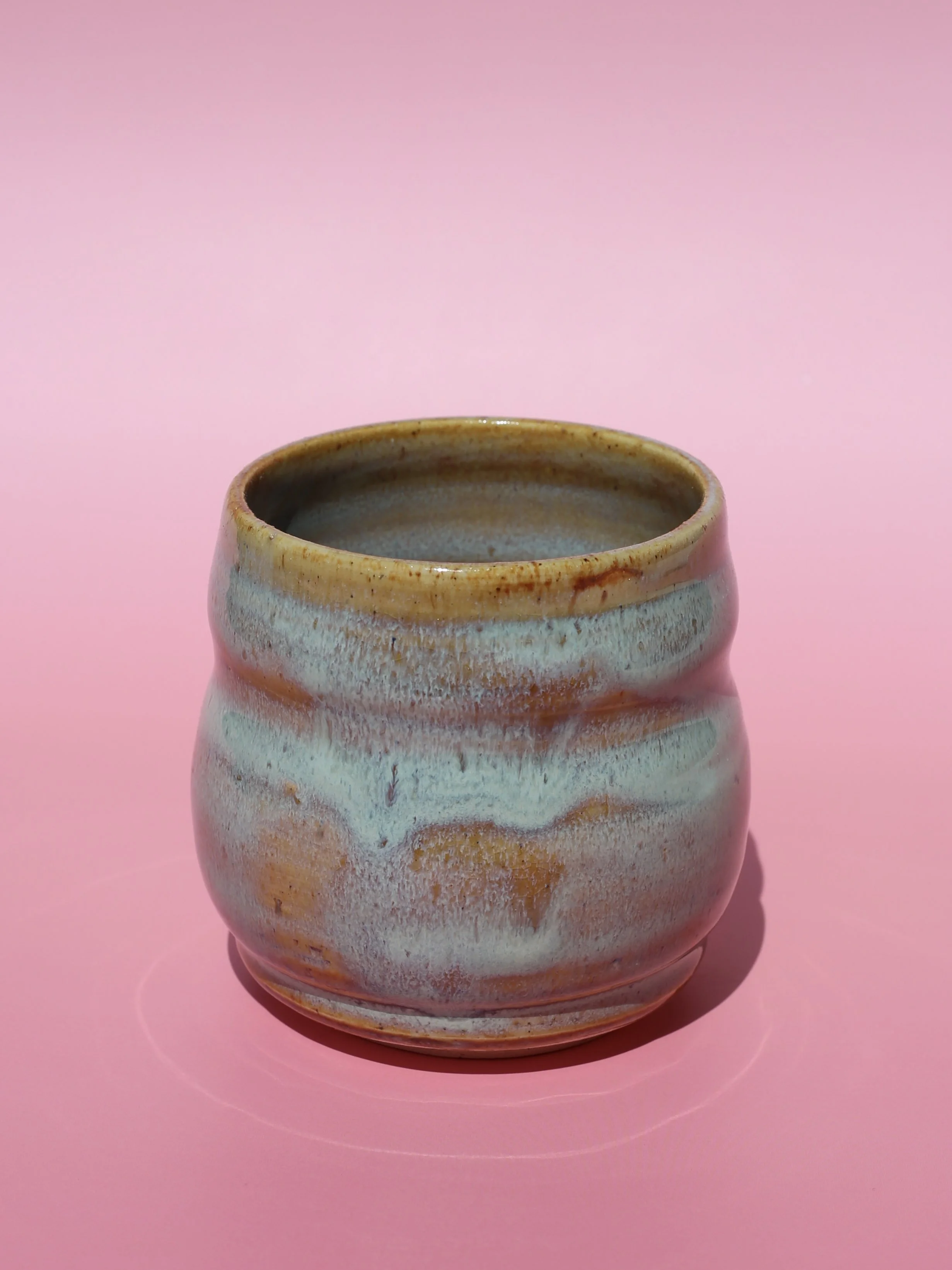 Ceramic vase with a textured, glazed surface in shades of beige, brown, and gray, placed on a pink background.