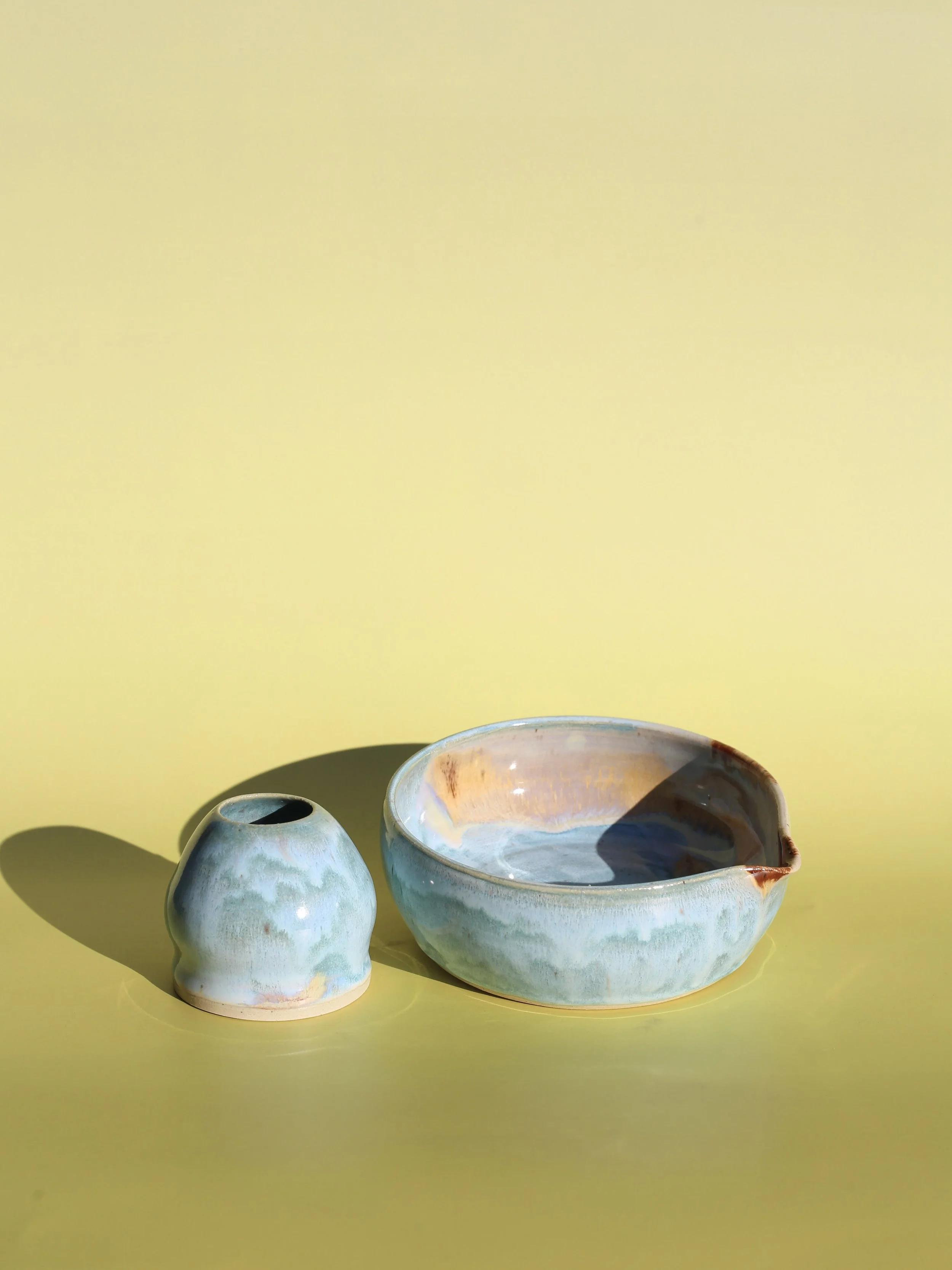 Two ceramic vessels with a light blue glaze, set against a yellow background. The vessels include a matcha whisk chasen holder and one larger bowl with a spout on the rim for pouring matcha or other liquids.