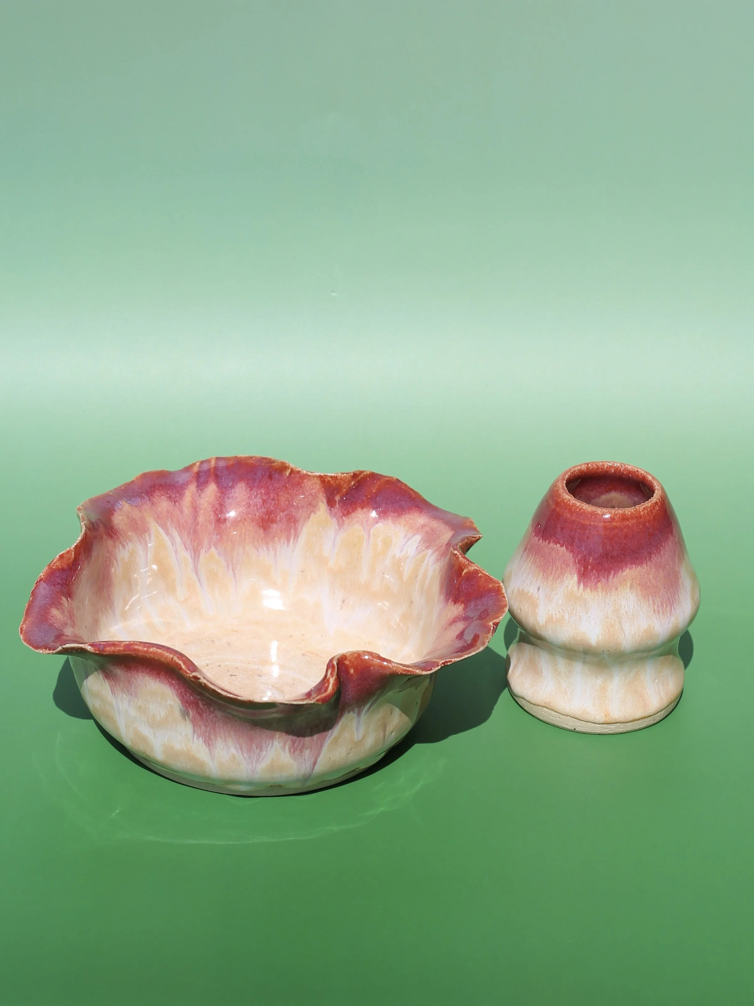 A large pink and cream-colored seashell bowl with a wavy, scalloped edge, sitting on a green surface. A smaller, similarly colored seashell with a rounded opening is positioned next to the bowl against a solid green background.