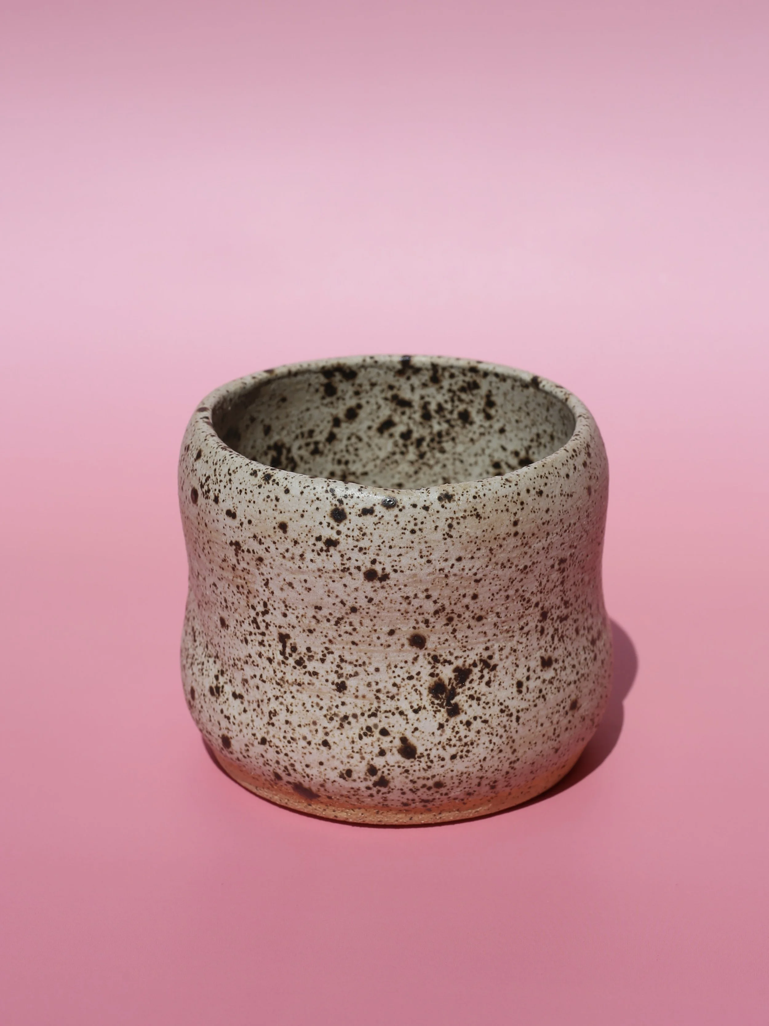 A handmade neutral speckled ceramic cup or vase against a pink background.