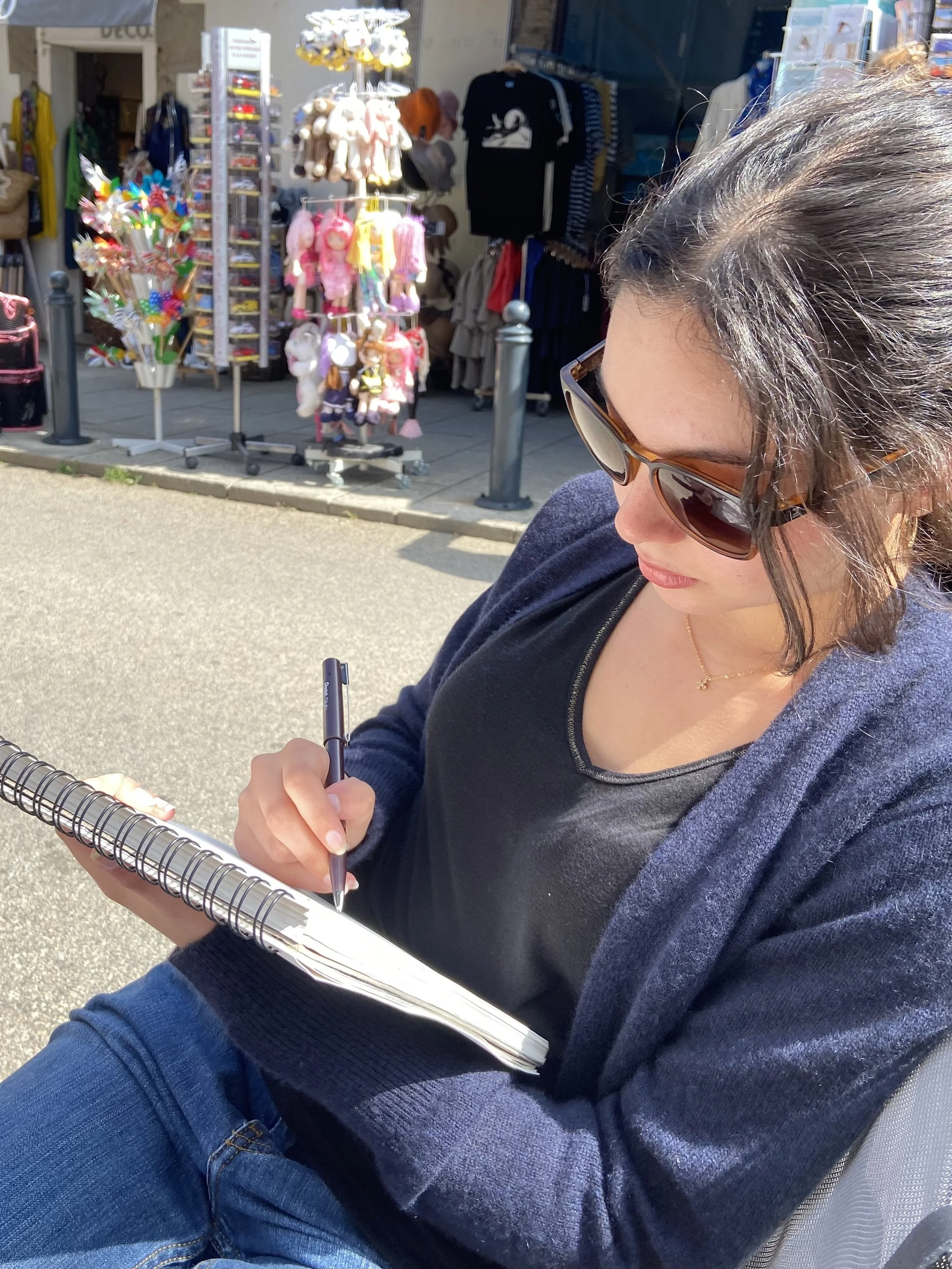 A woman with dark hair wearing sunglasses and a black top, sitting outdoors and writing in a spiral notebook. Behind her, there is a storefront with colorful toys, plush animals, and clothing items on display.
