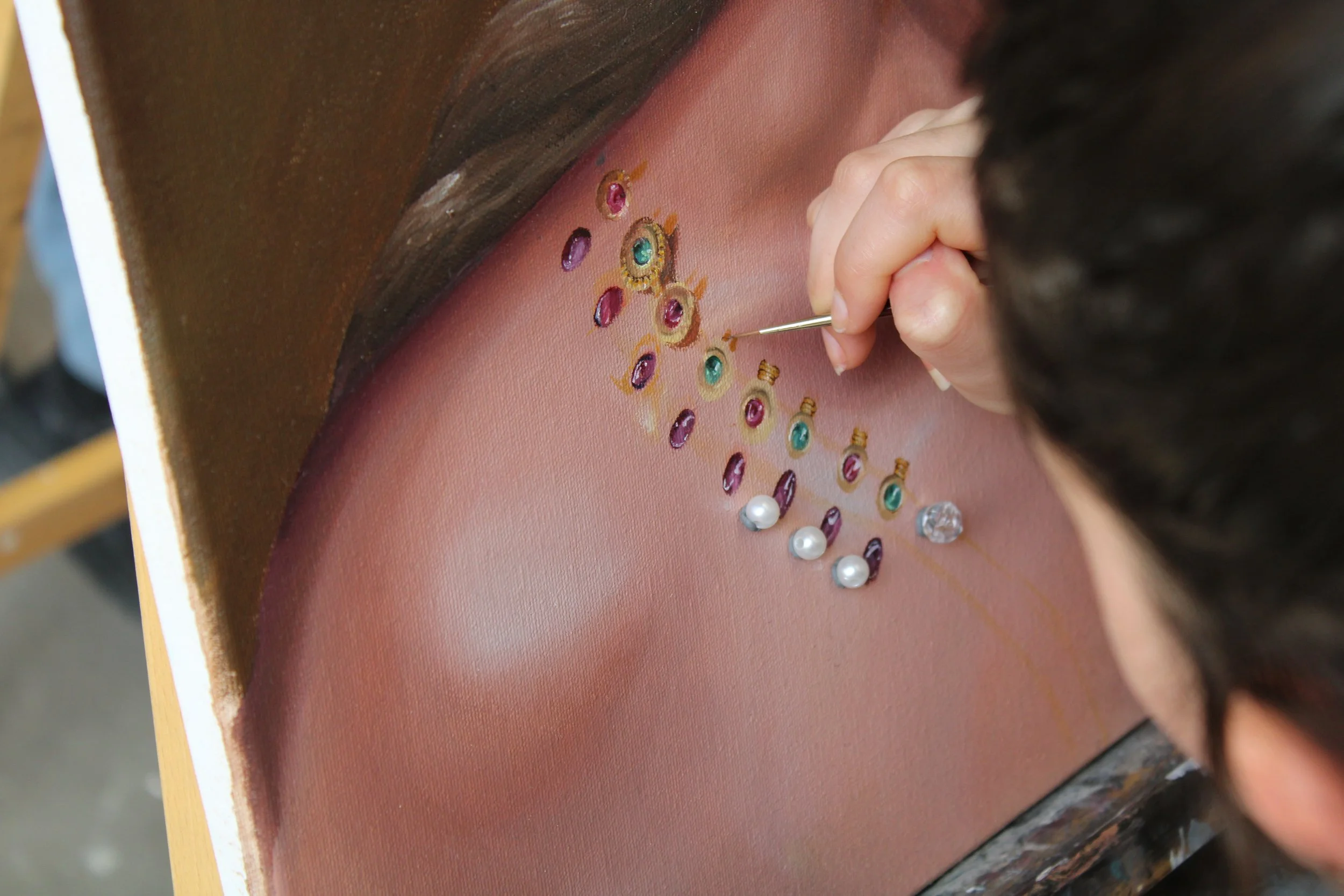 Close-up of an artist painting a detailed portrait of a woman with dark hair, using a fine brush to add embellishments and jewelry details on the skin.
