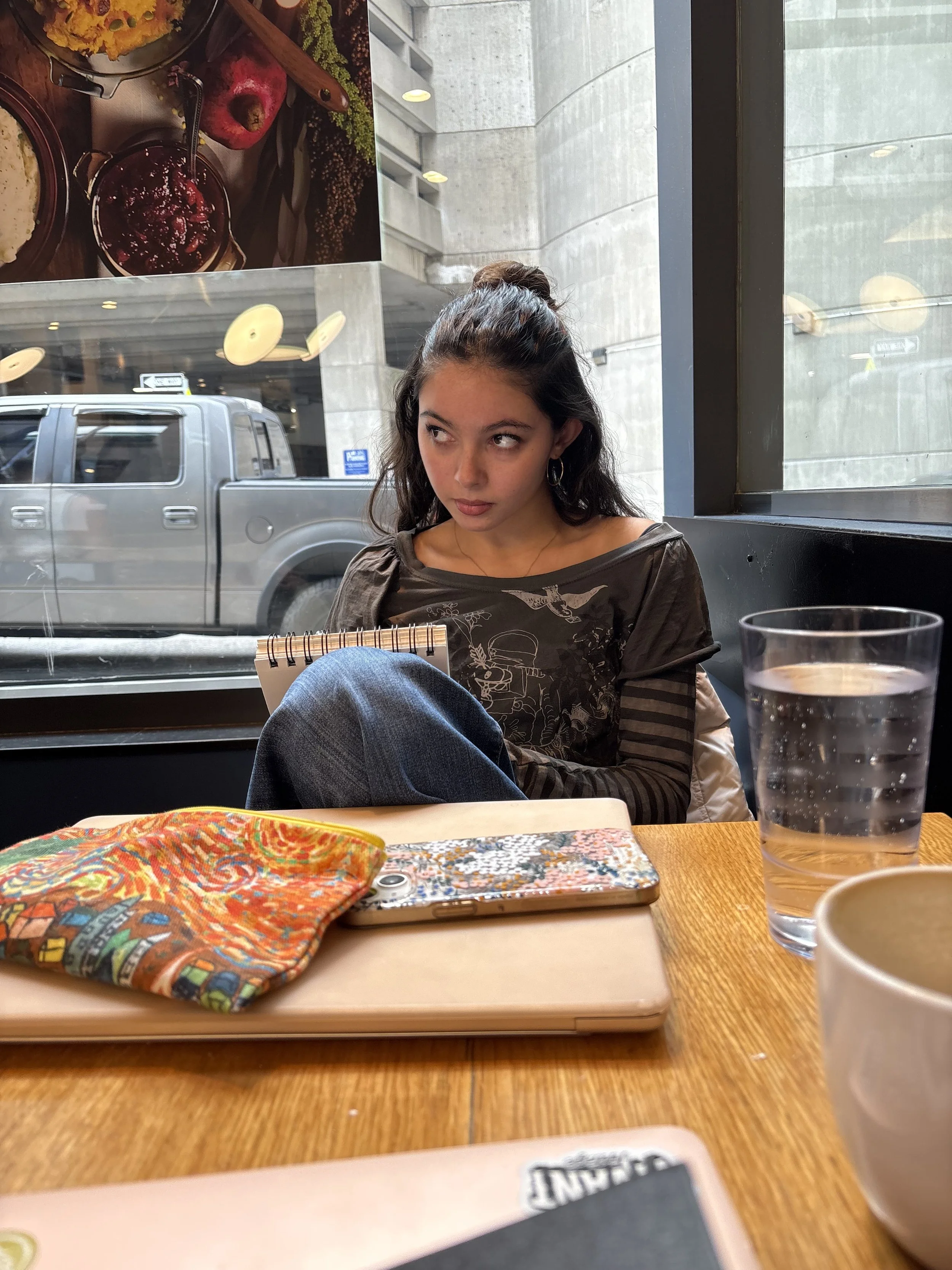 A young woman with dark, curly hair tied back in a bun, sitting at a restaurant table, looking to the side. There is a glass of water and a colorful cloth napkin on the table, with a patterned phone case and part of a menu visible. The background shows a large window, a gray building outside, and a parked truck.