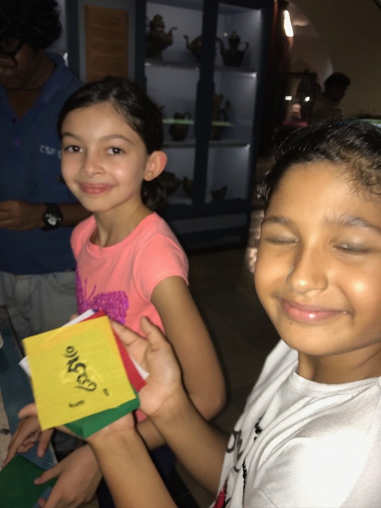 Two smiling children, a girl in a pink shirt and a boy in a white shirt, are holding colorful cards or papers inside a room with shelves of decorative items in the background.