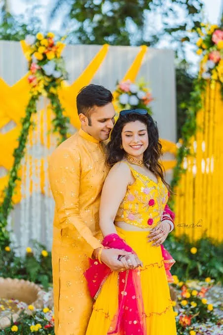 A young couple dressed in traditional yellow and pink clothing, celebrating a festive occasion outdoors with colorful floral decorations and a bright yellow background.