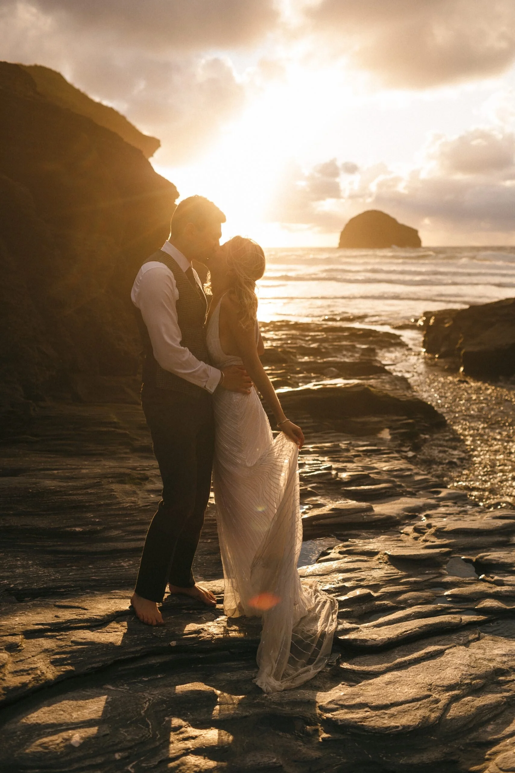 A couple sharing a romantic moment at the beach during sunset, standing close and touching foreheads, with a large rock formation in the ocean in the background.