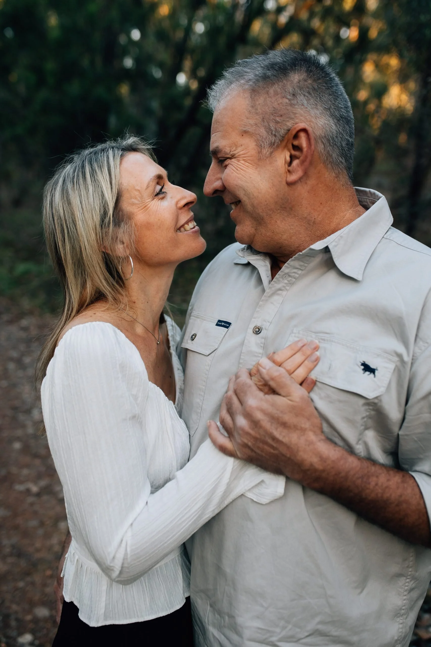 A middle-aged couple stands outdoors, smiling and looking into each other's eyes, holding hands, surrounded by trees.