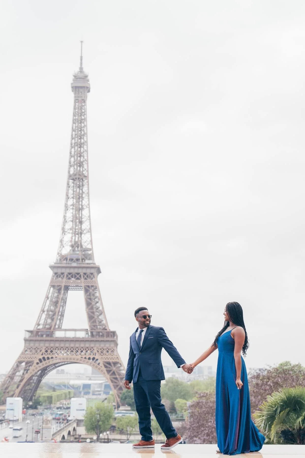 A couple holding hands and dancing in front of the Eiffel Tower on a cloudy day.
