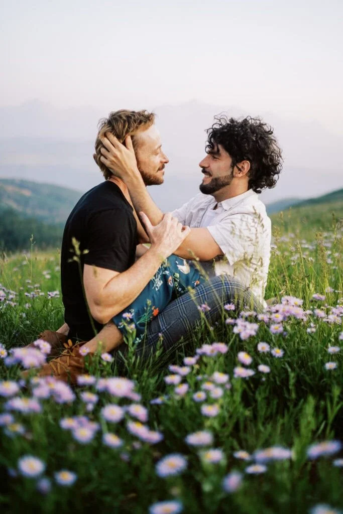 Two men sitting in a field of flowers, facing each other, holding each other's faces, sharing an intimate moment outdoors with mountains in the background.