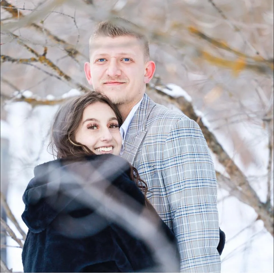 A smiling young couple standing outdoors in a snowy winter forest, embracing each other and looking at the camera.