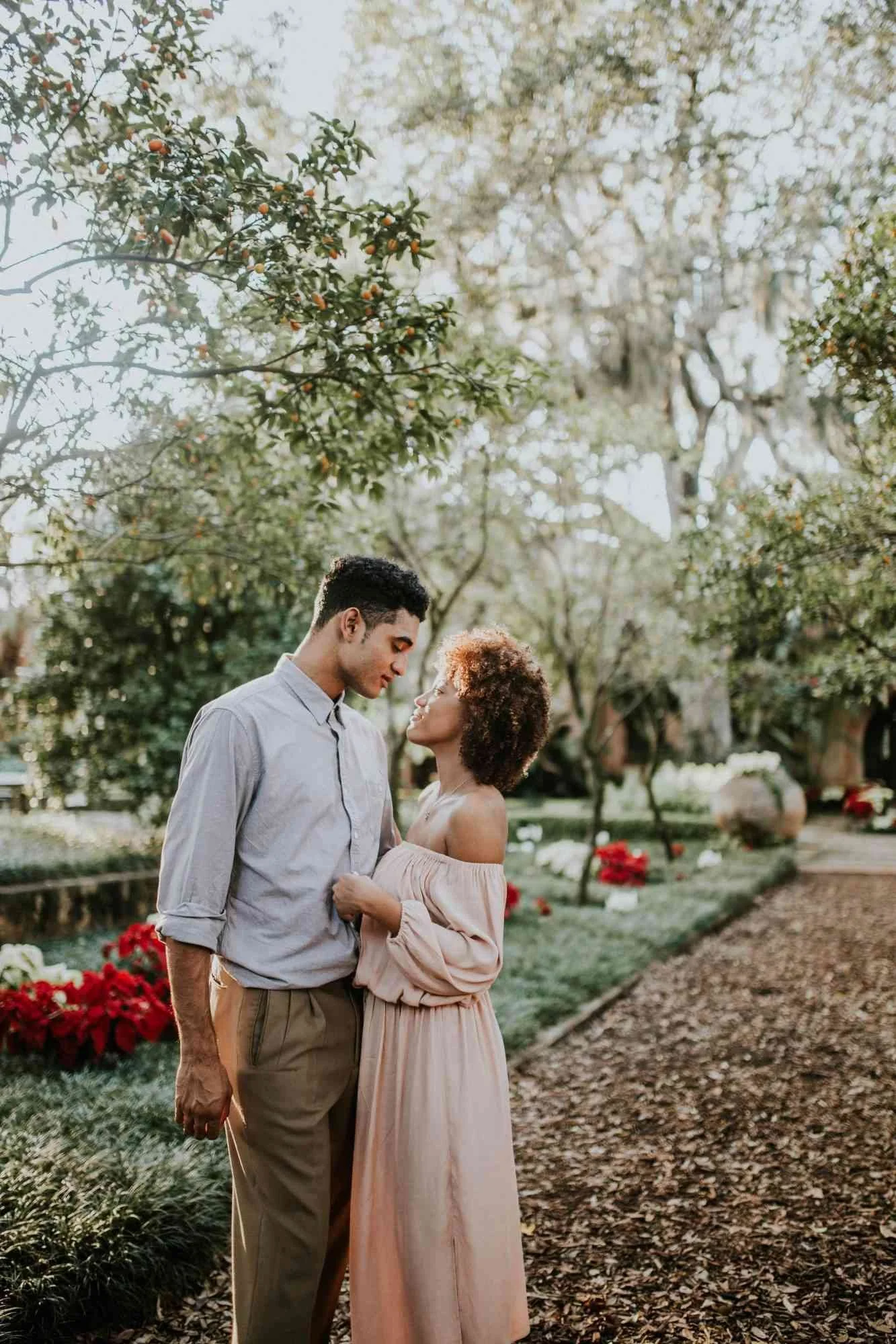 A man and a woman standing closely together outdoors in a park with trees. The man is wearing a light gray shirt and tan pants, and the woman is wearing a long, off-the-shoulder pink dress. They are gazing at each other lovingly. There are red flowers and a pathway visible in the background.