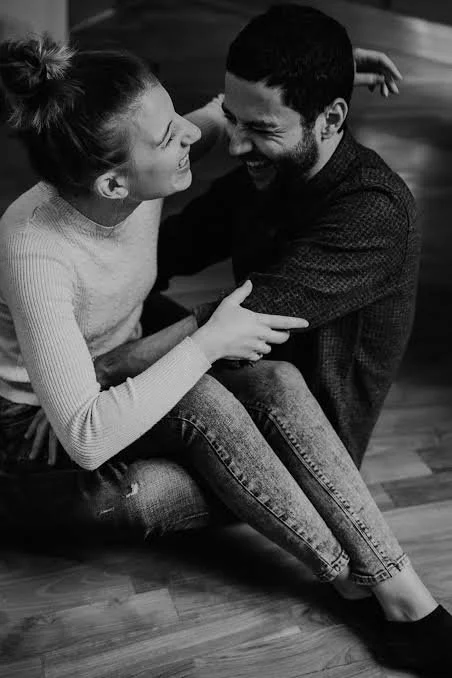 A smiling young woman and man sitting on a wooden floor, holding each other and laughing together.