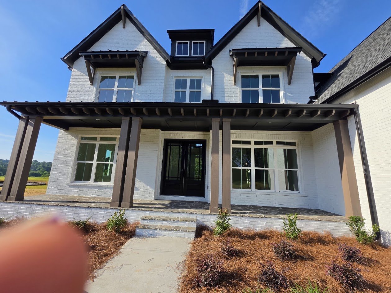 Modern three-story house with white brick exterior, multiple windows, and a black front door, surrounded by landscaping with small plants and a concrete walkway.