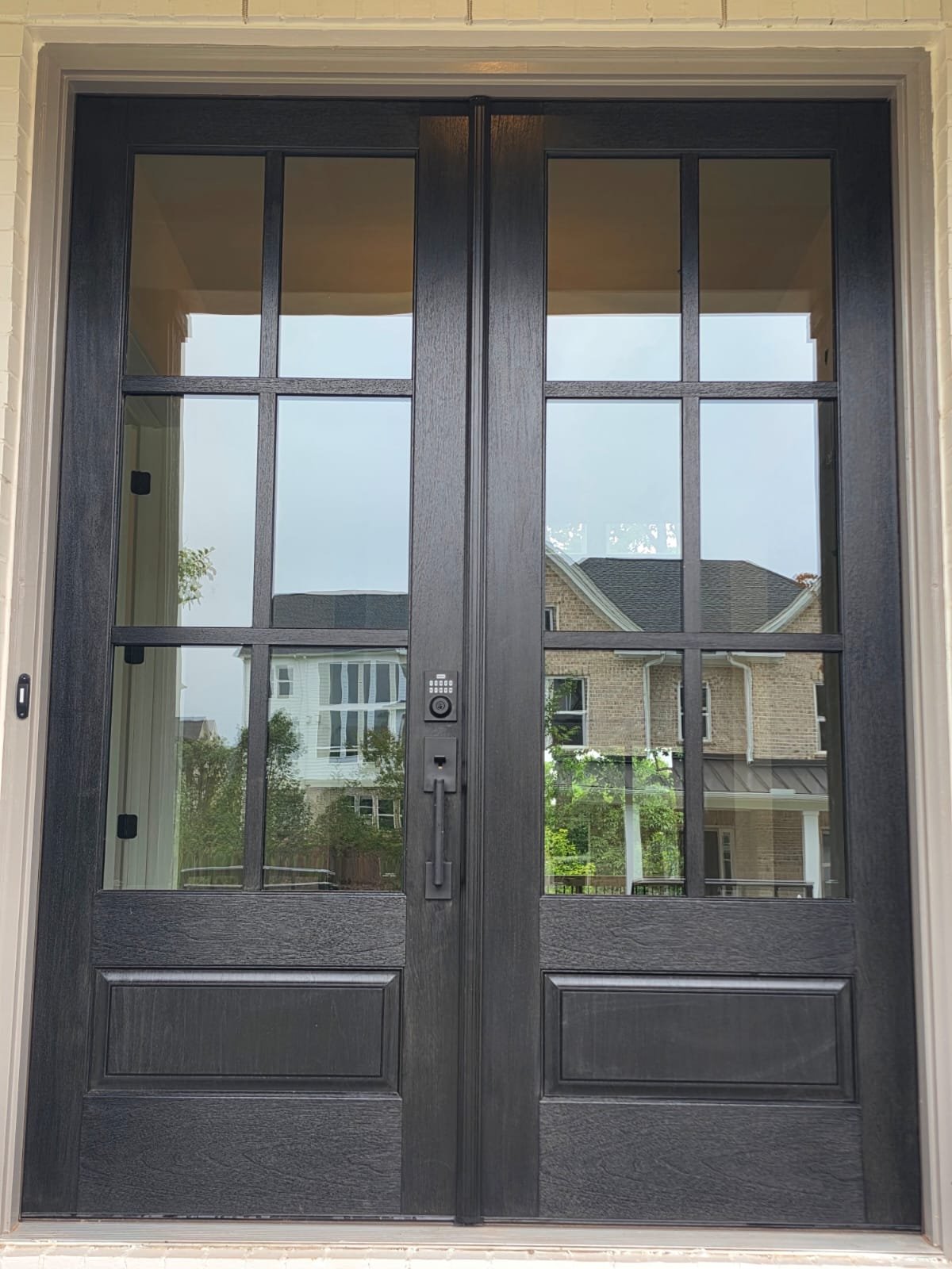 Black double front door with glass panels and a keypad lock, reflecting neighboring houses and trees.