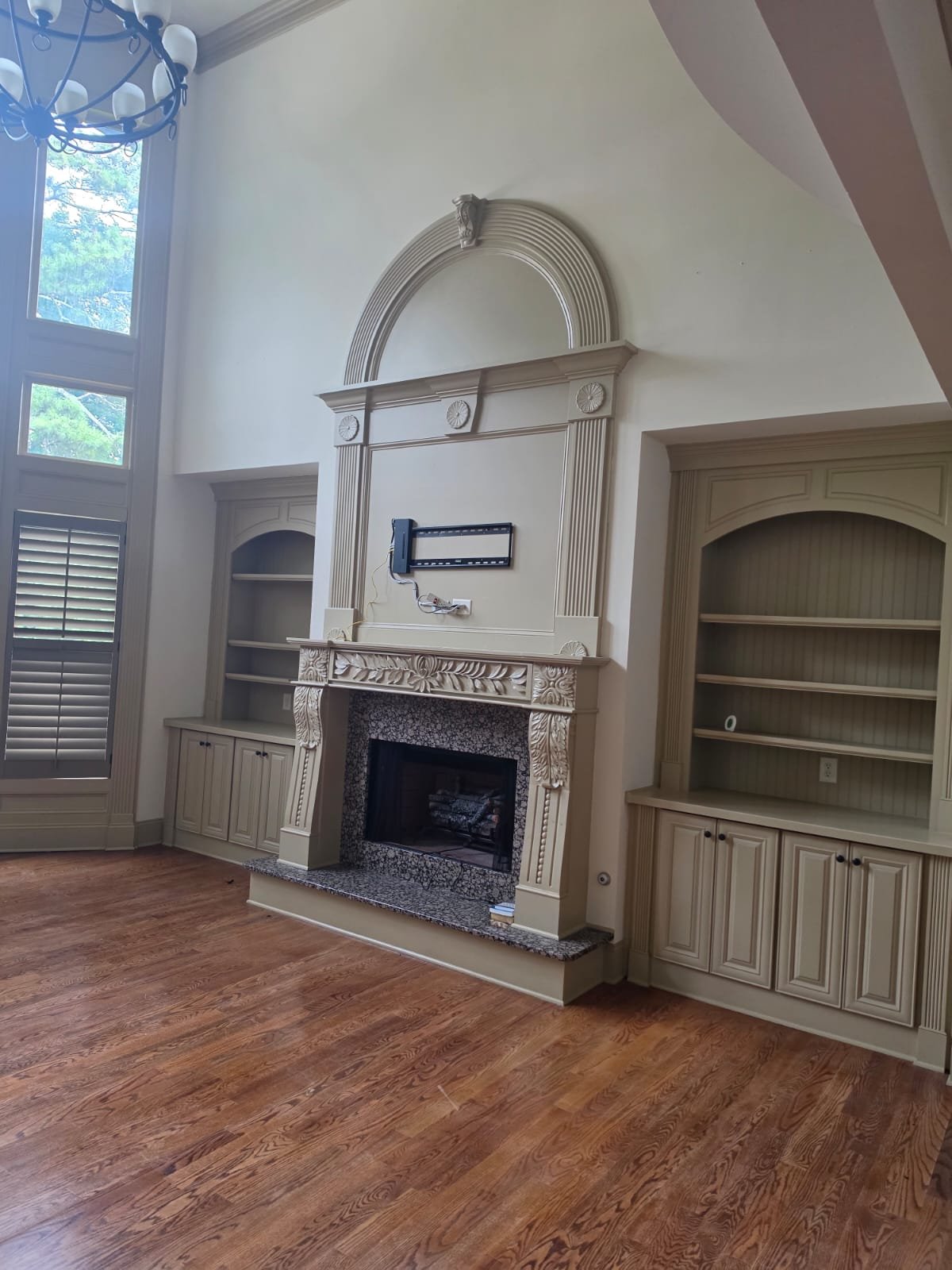 Living room with wood flooring, built-in bookshelves, ornate fireplace, and large window with plantation shutters, chandelier hanging from ceiling.