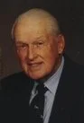 A portrait of an elderly man with white hair, wearing a dark suit, white shirt, and tie, smiling gently.