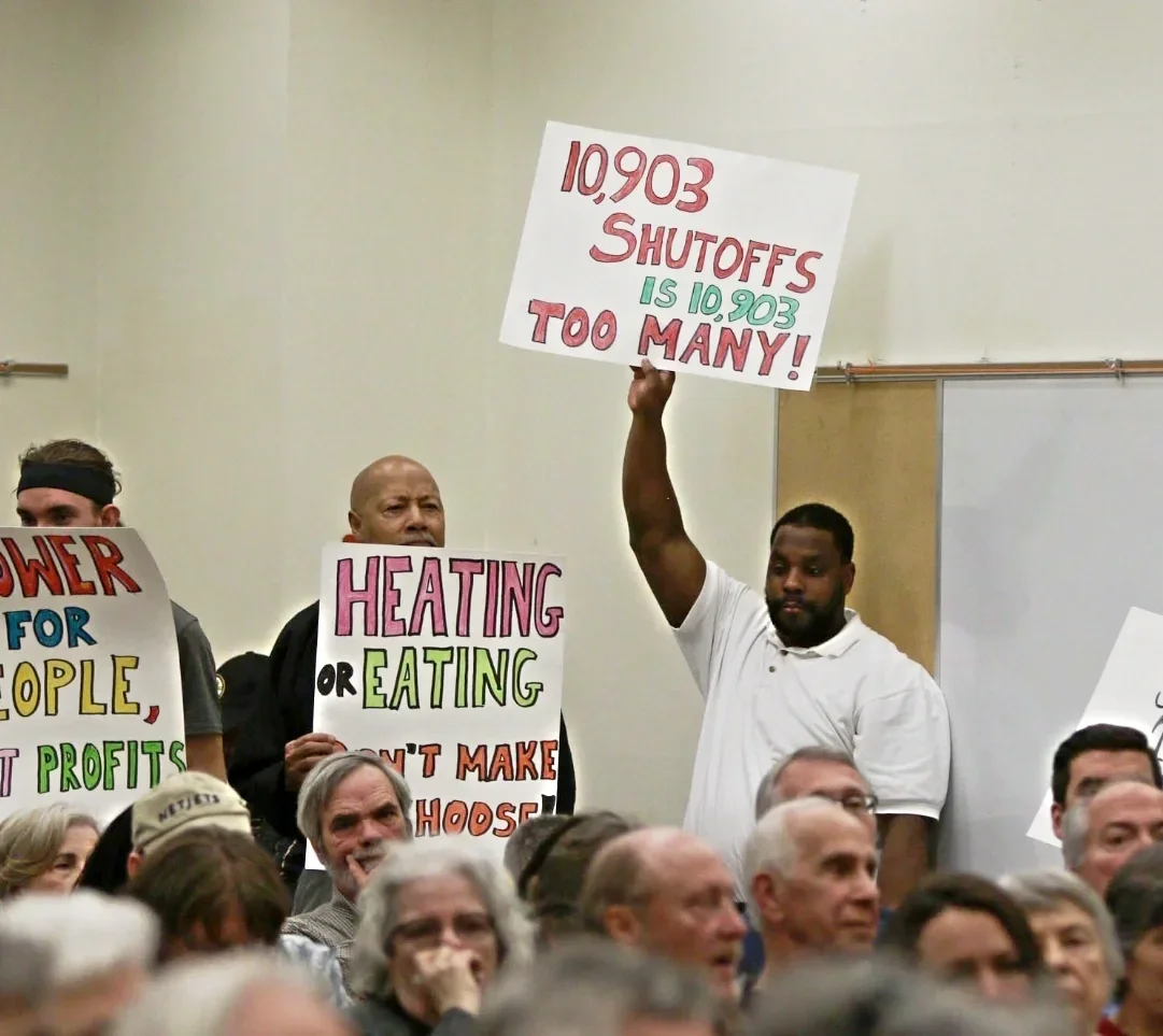 A man at a utility rate hike public hearing holding up a sign that says "10,903 Shut Offs is 10,903 Too Many", next to a man with a sign that says "Heating or Eating, Don't Make Us Choose"