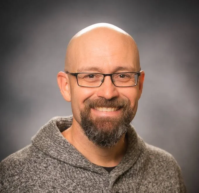 A smiling man with a bald head, glasses, a beard, and a mustache, wearing a gray sweater against a gray background.