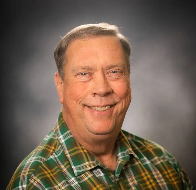 A man with light skin, short light brown hair, smiling, wearing a green plaid shirt, against a dark gray background.