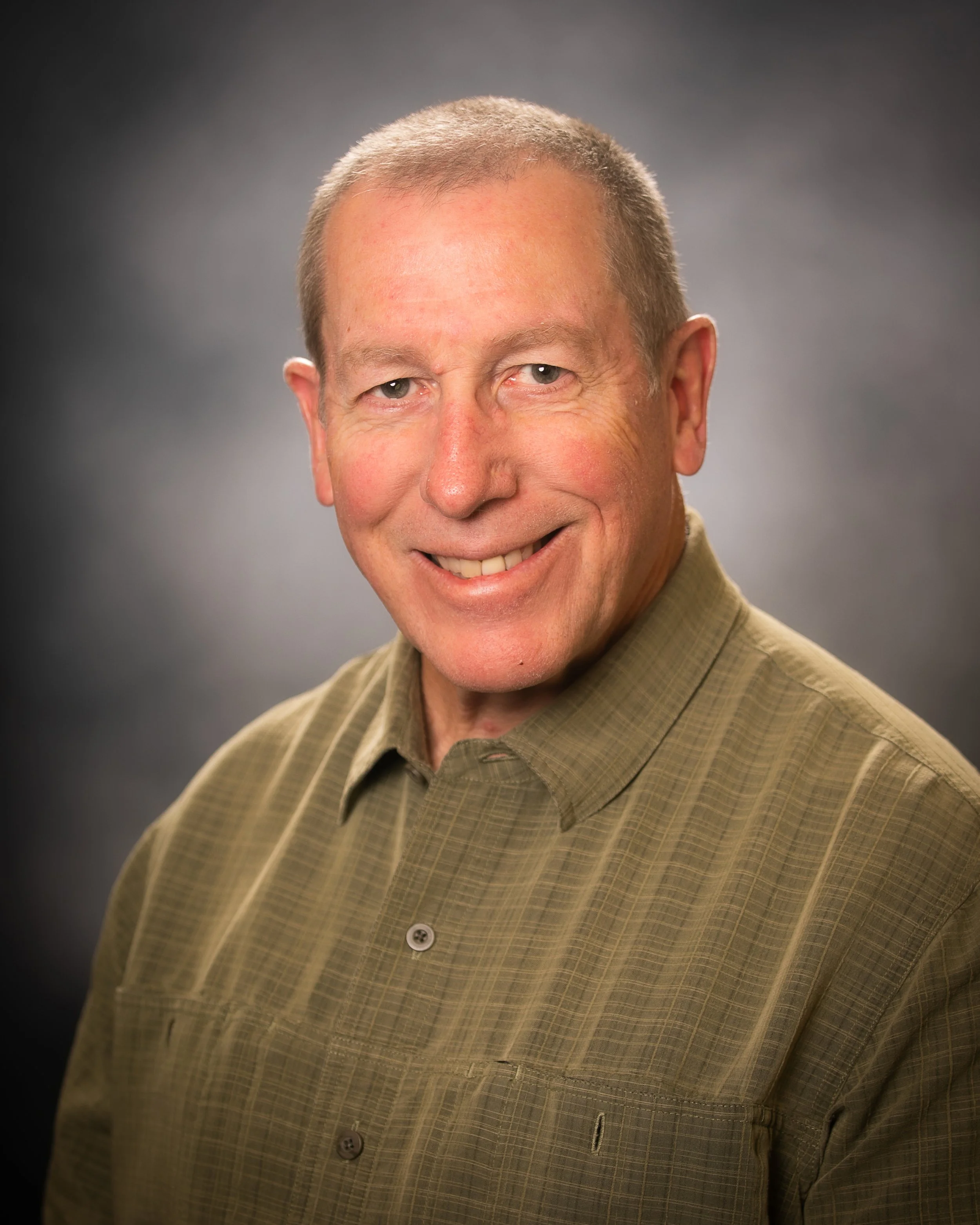 Close-up of a smiling middle-aged man with short hair, wearing a tan button-up shirt, against a dark gray background.