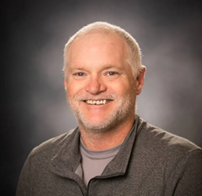 Portrait of a smiling man with short white hair and beard, wearing a gray zip-up jacket against a dark blurred background.
