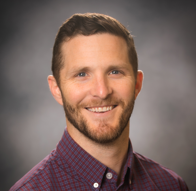 A smiling man with a beard and short brown hair, wearing a red checkered shirt, on a gray background.