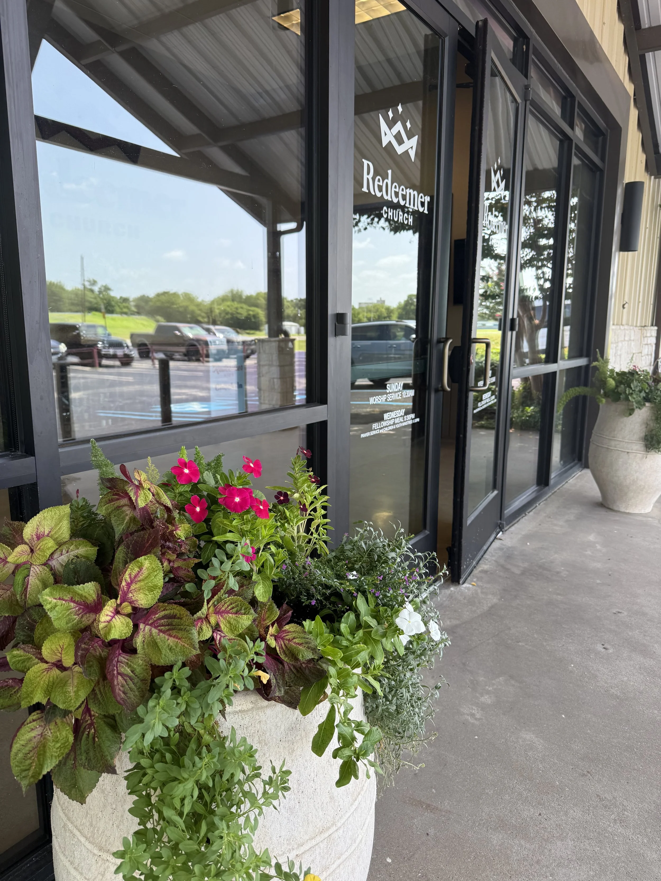 Entrance door of Redeemer Church with glass windows, surrounded by flower pots with pink, white, and green plants, sidewalk, parking lot, and trees outside.