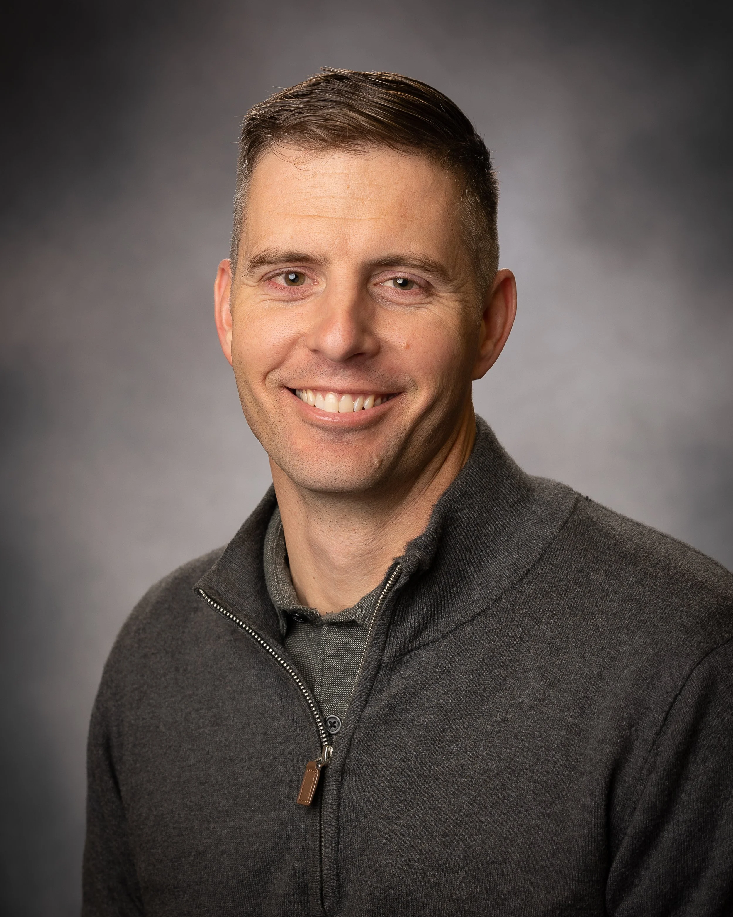 Smiling man with short brown hair, wearing a gray zip-up sweater over a gray shirt, against a dark gray background.
