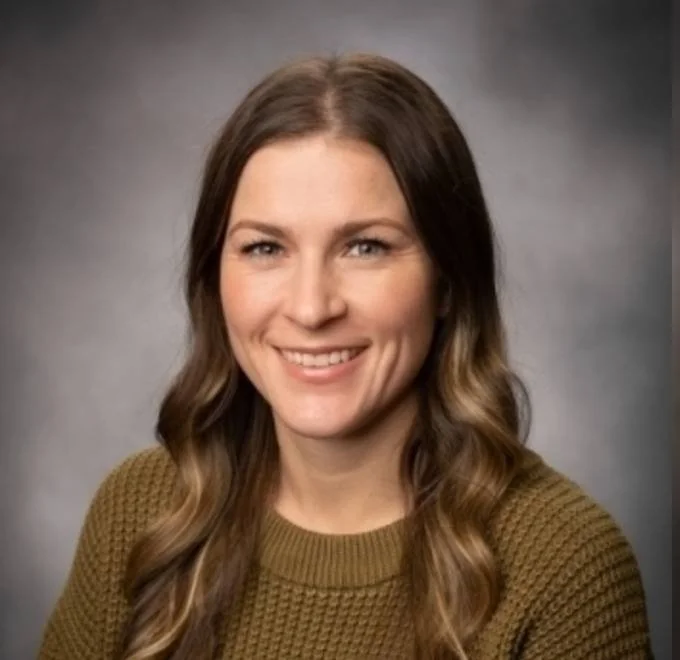 A woman with long wavy brown hair smiling, wearing a brown knit sweater, against a gray gradient background.