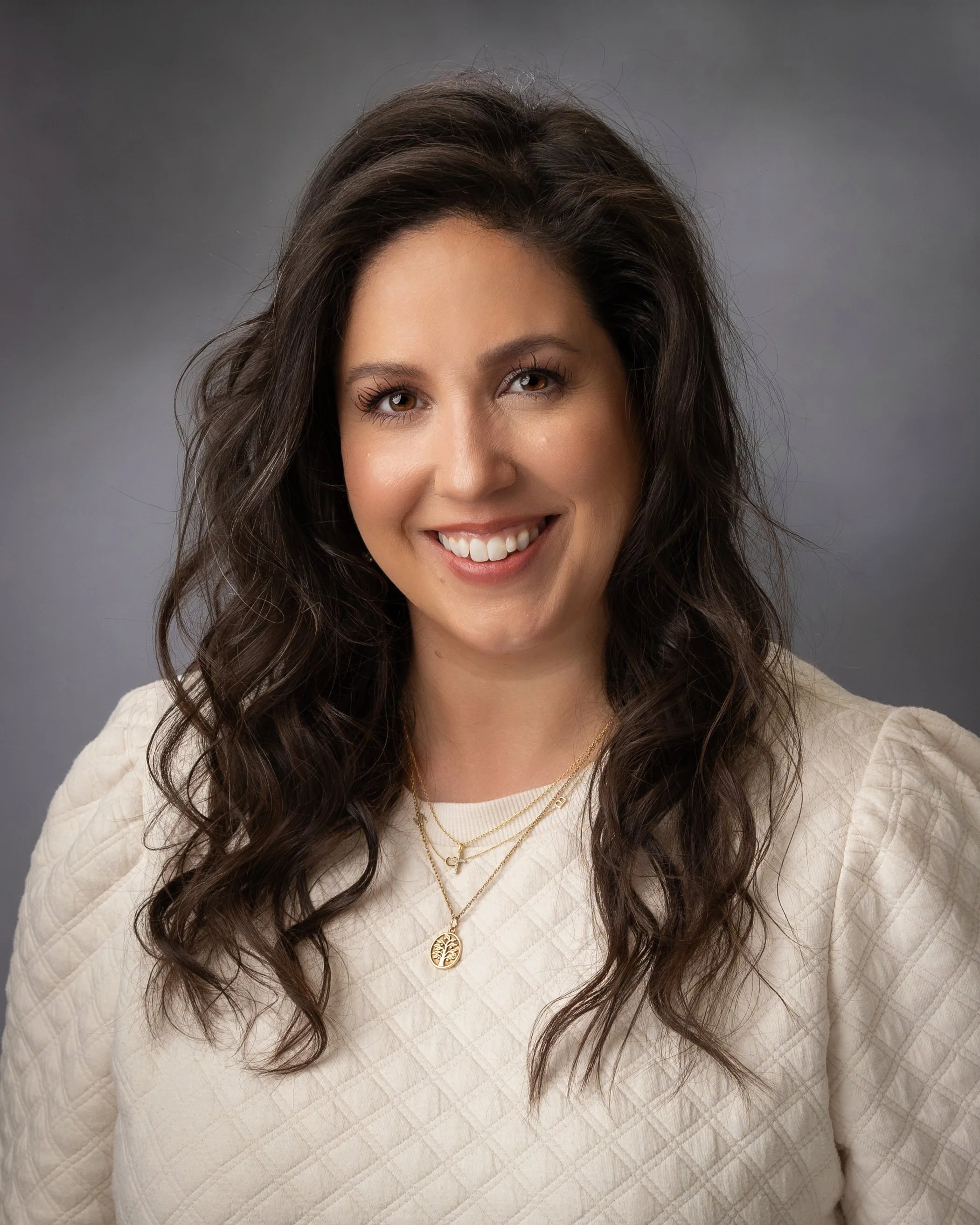 A smiling woman with long, wavy dark hair wearing a beige diamond-patterned top and layered gold necklaces, posing against a gray background.