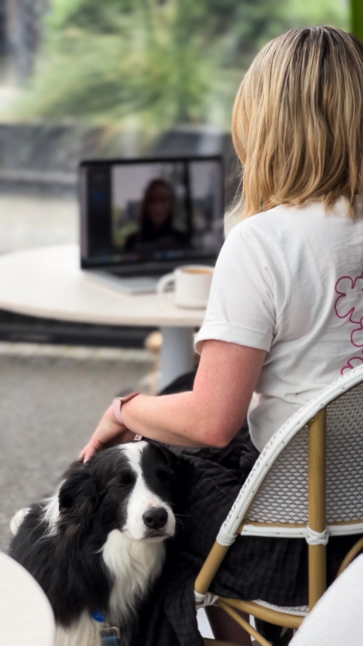 A person with blonde hair sitting on a white outdoor chair next to a black and white dog, with a laptop and coffee cup on a white table in front of them, outdoors with greenery in the background.