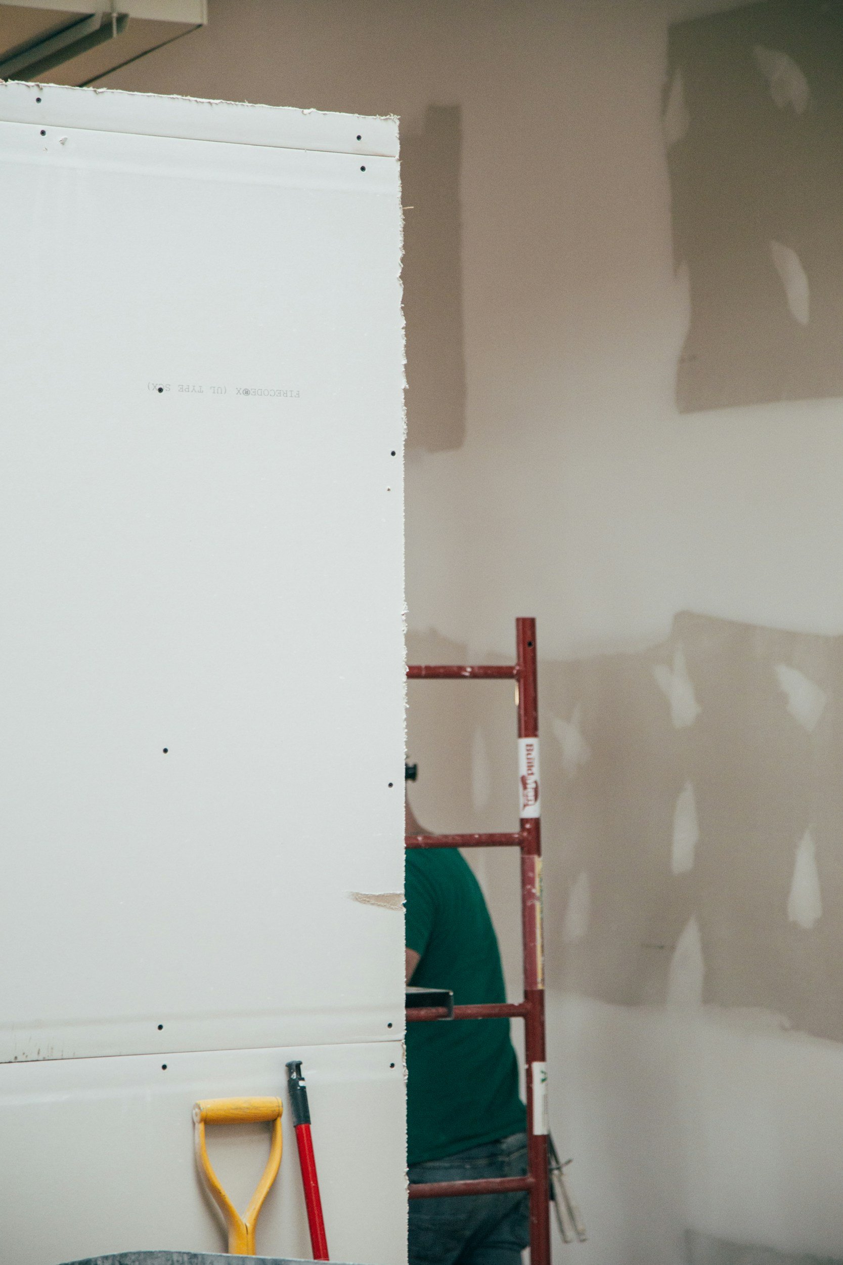 Construction worker wearing a green shirt and jeans standing behind a metal frame, partially obscured by a white board, on a construction site with unfinished walls and tools.
